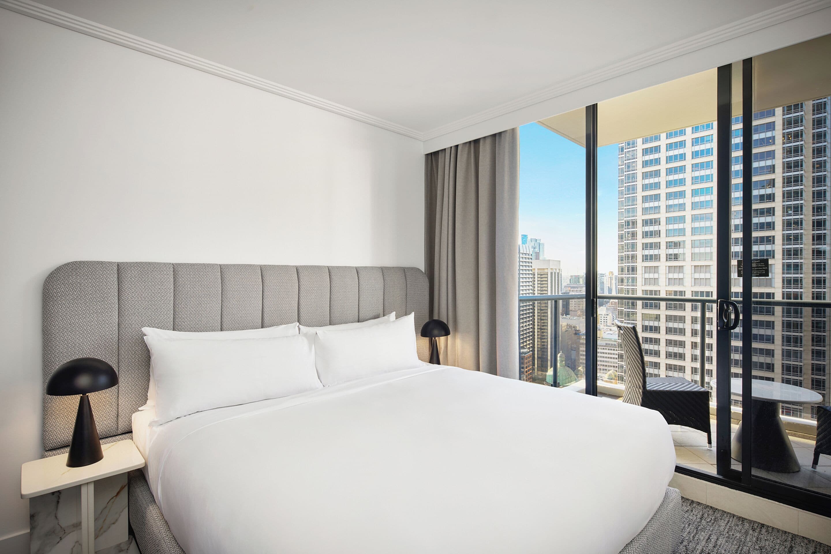 A modern hotel room with a large bed, a nightstand, a lamp, and a view of the city through floor-to-ceiling windows.