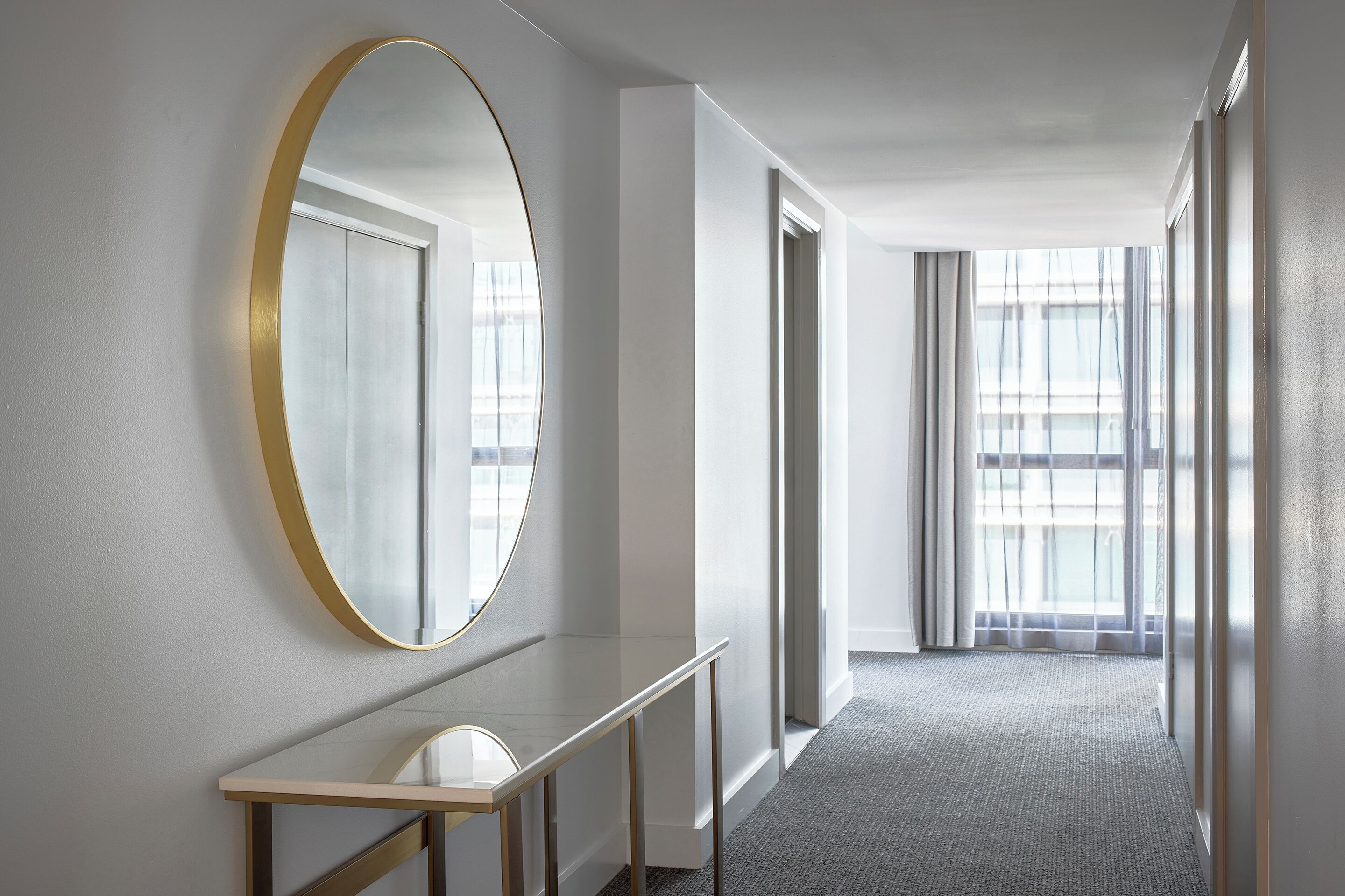 A hallway with a large mirror, a console table, and a carpeted floor leading to a room with large windows.
