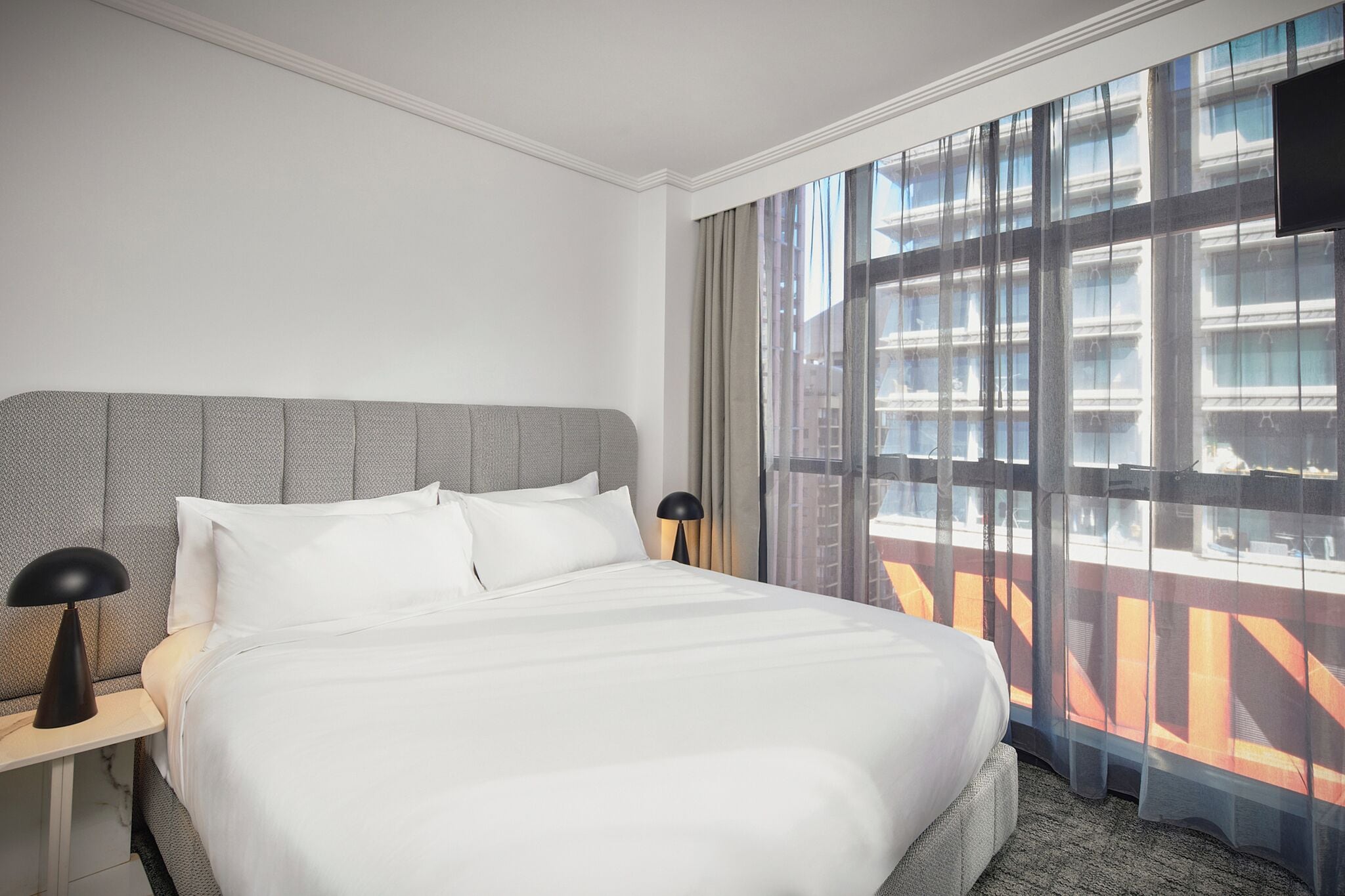 A hotel room with a large bed, a nightstand with a lamp, and a view of a building through sheer curtains.