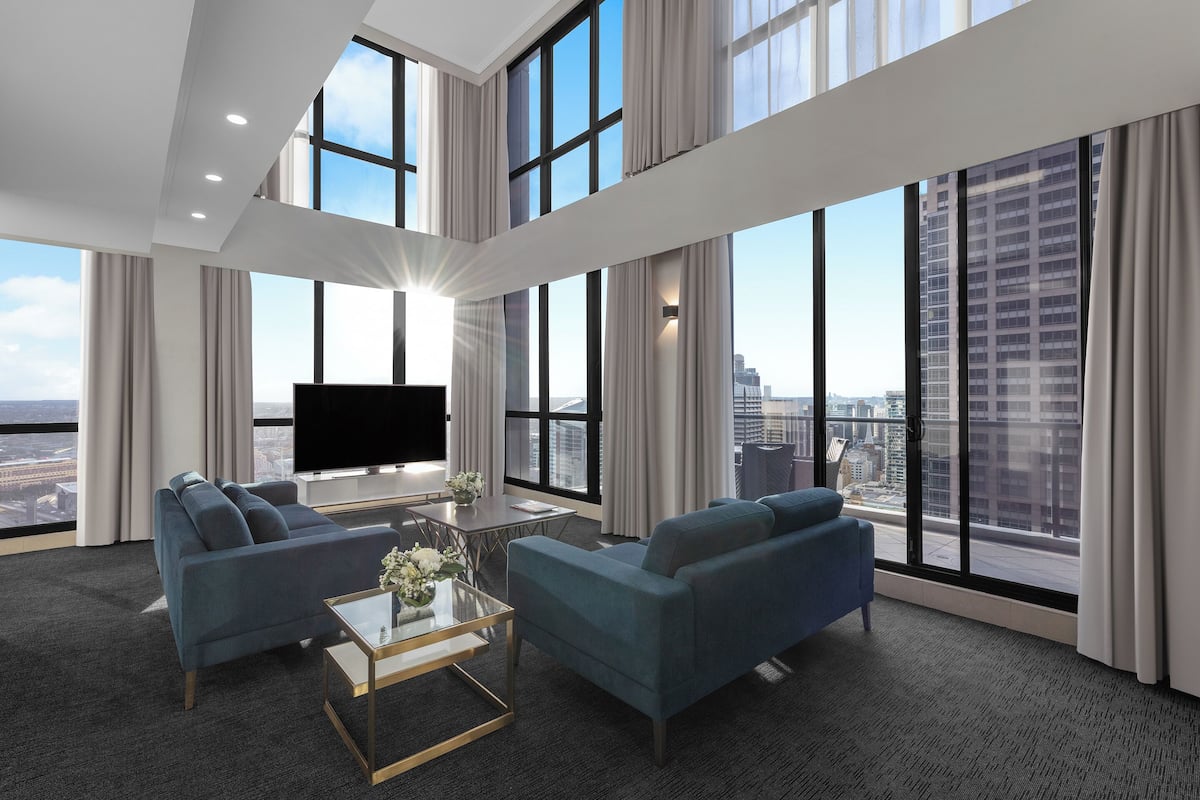 A modern hotel room with a large TV, two sofas, a coffee table, and a city view.