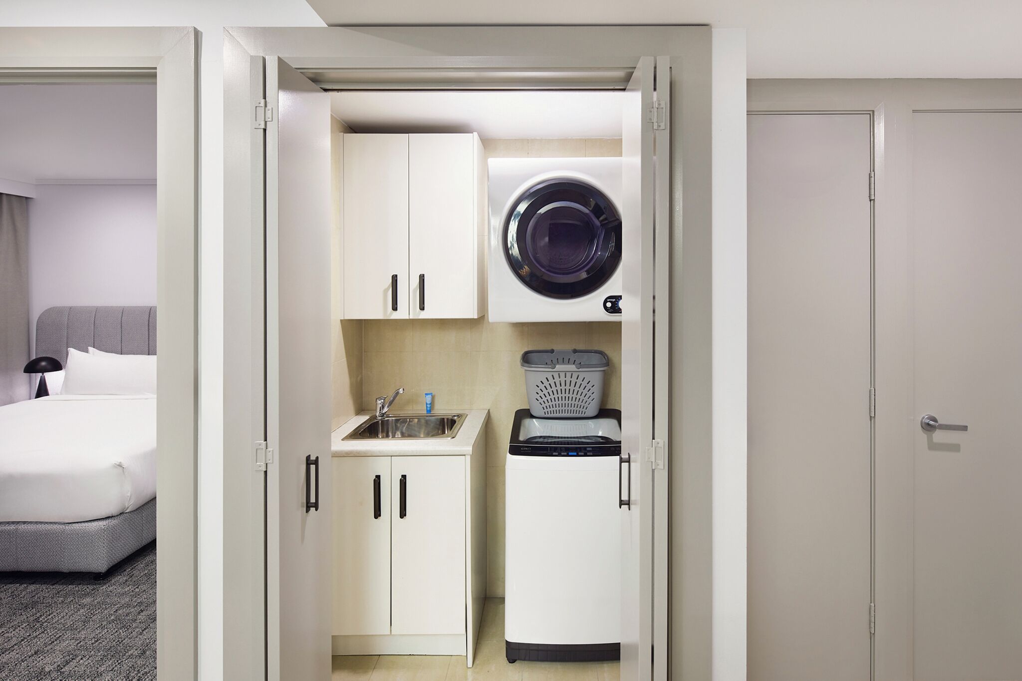 A compact hotel room with a bed, a kitchenette with a washing machine, and a small sink.