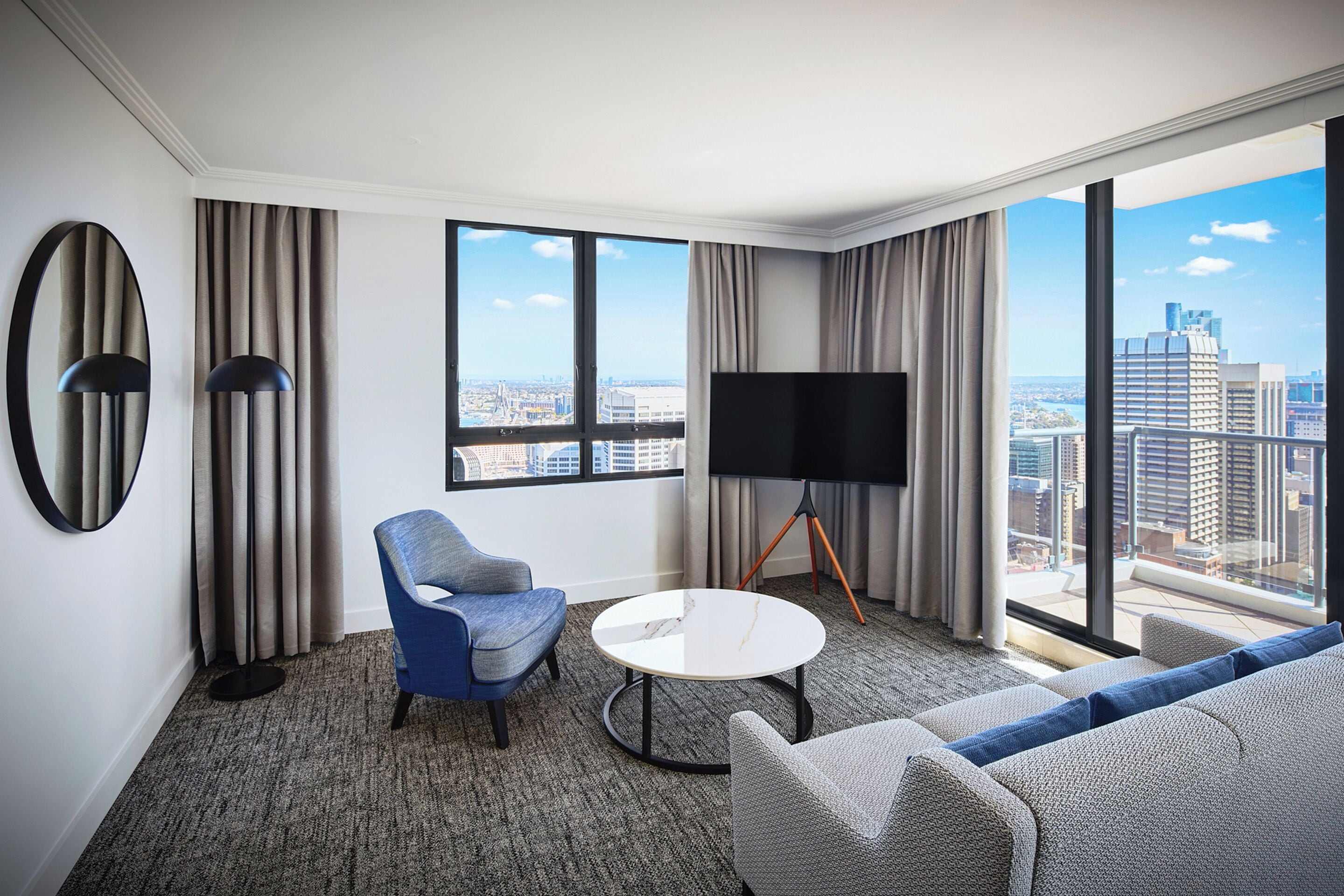 A modern hotel room with a large window, a flat-screen TV, a round coffee table, and a sofa.