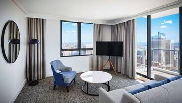 A modern hotel room with a large window, a flat-screen TV, a round coffee table, and a sofa.