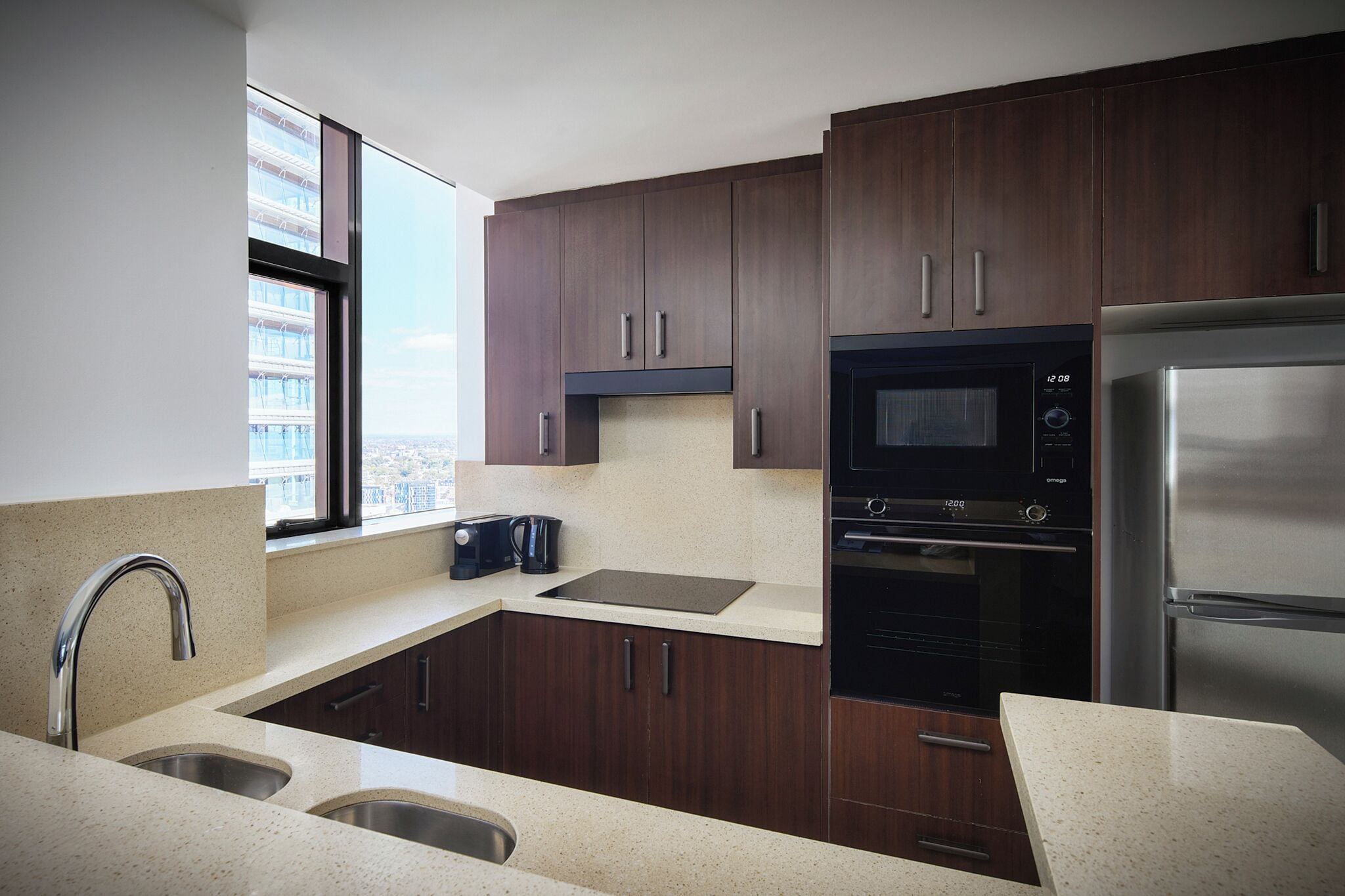 A modern kitchen with dark wooden cabinets, stainless steel appliances, and a large window offering a city view.