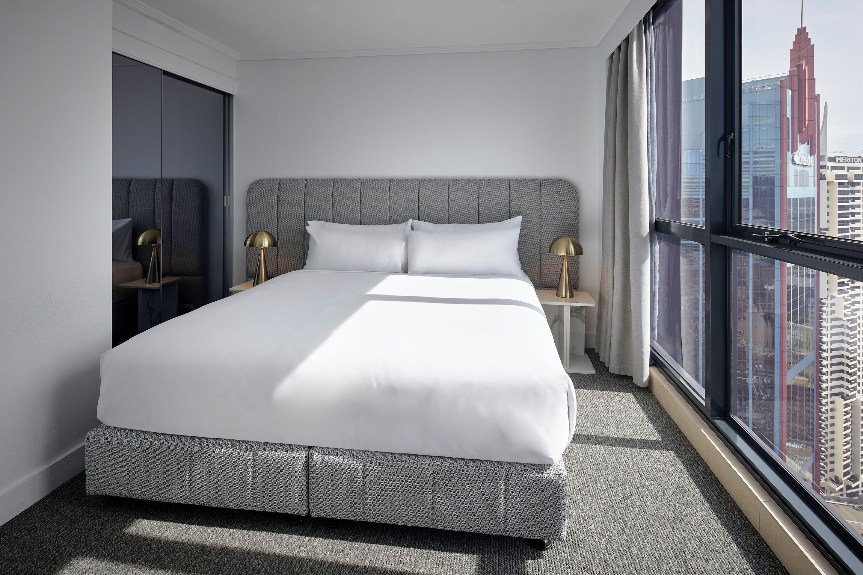 A modern hotel room with a large bed, two bedside tables with lamps, a view of a cityscape through a large window, and a city skyline visible outside the window.