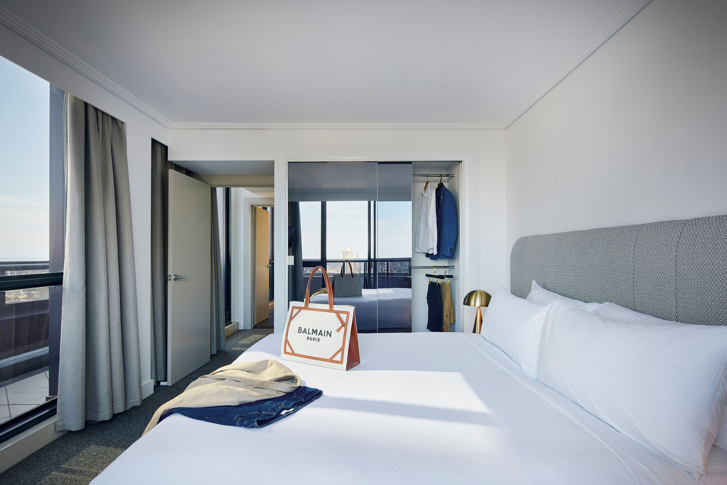 A hotel room with a large bed, a view of the city, a balcony, and a Balmain Paris handbag on the bed.