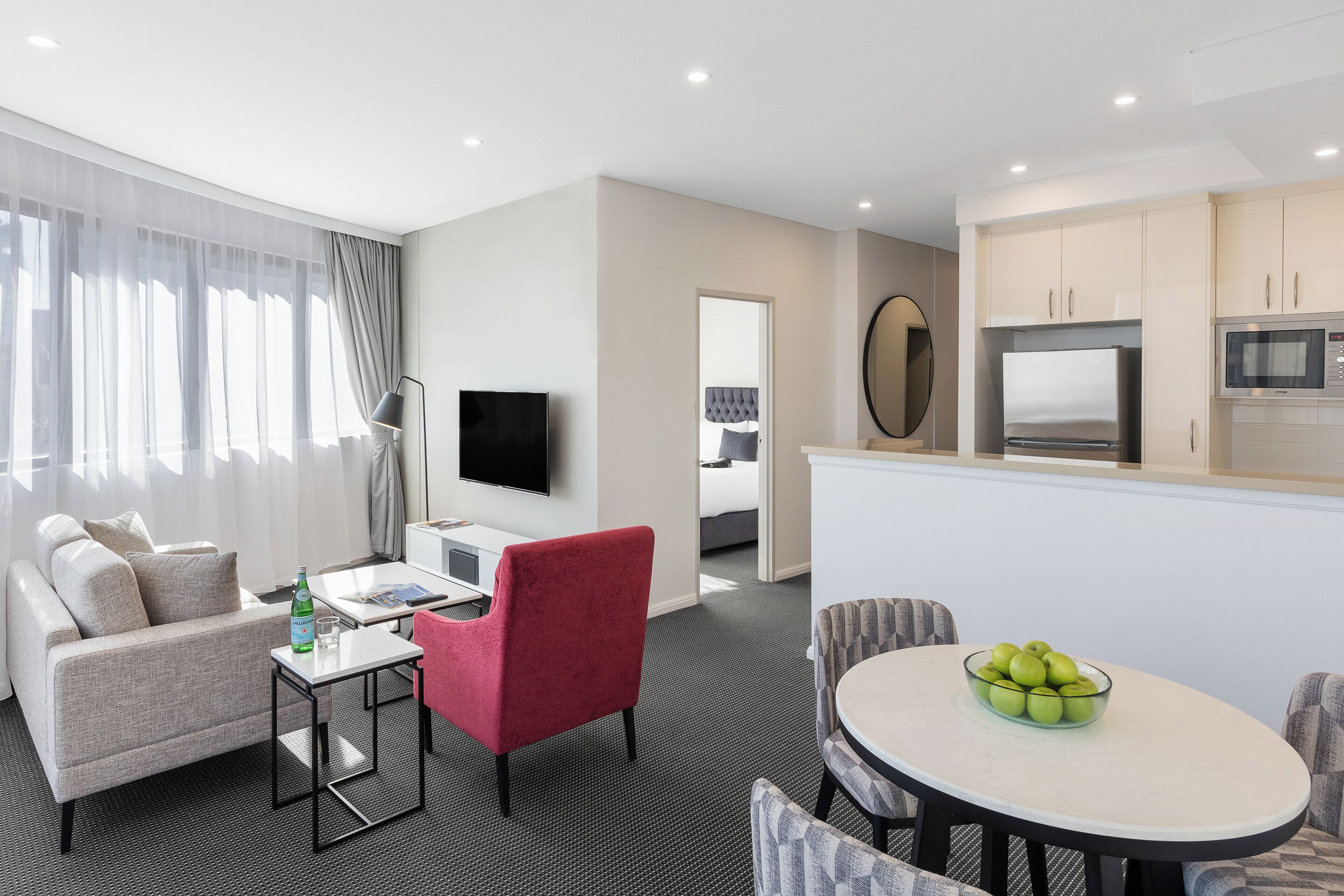 A hotel room with a sofa, a red chair, a dining table with apples, a TV, a kitchen area with a microwave, and a large window with curtains.