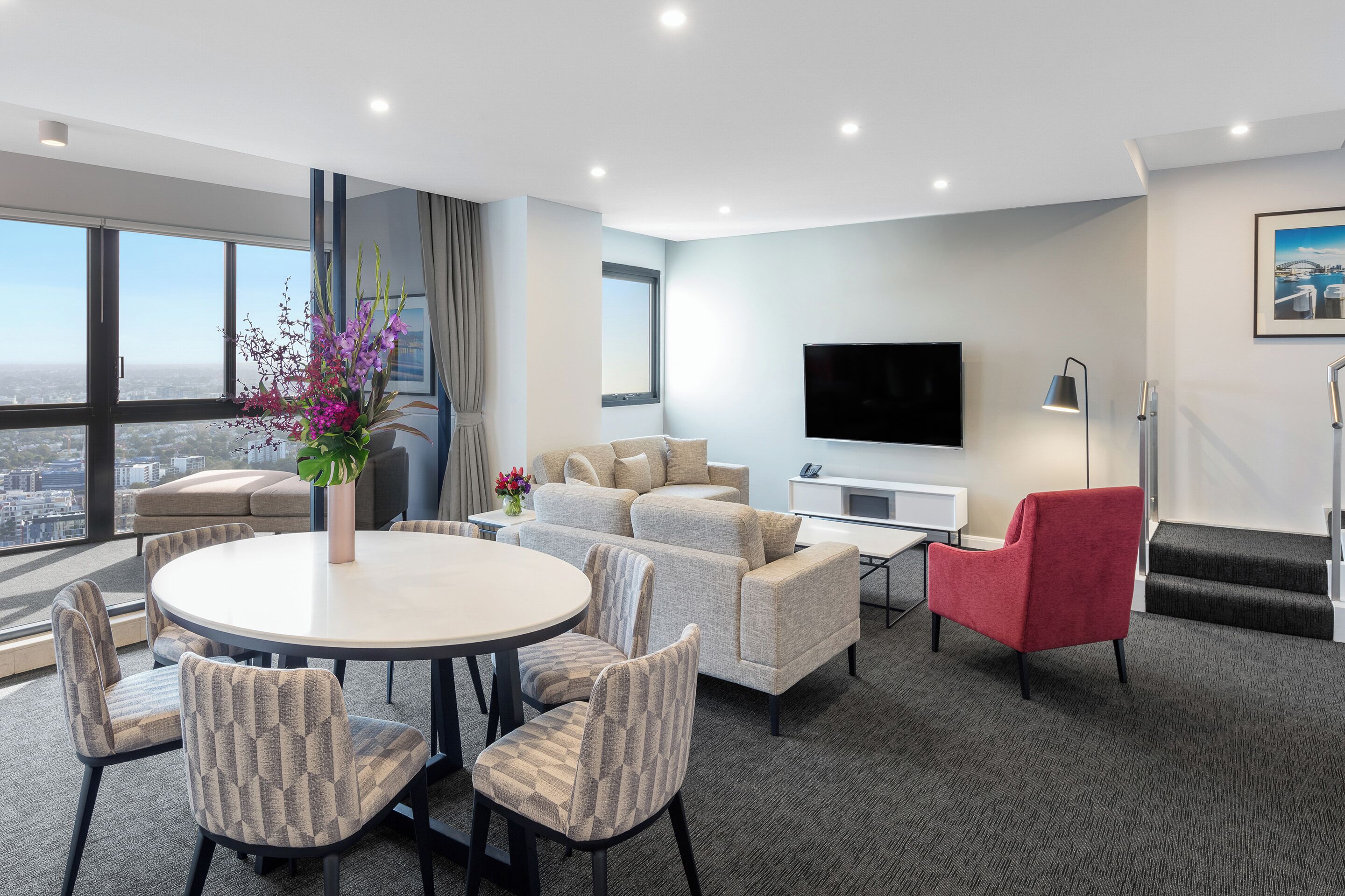A modern hotel room with a round dining table, cushioned seating, a flat-screen TV, and a large window offering a city view.