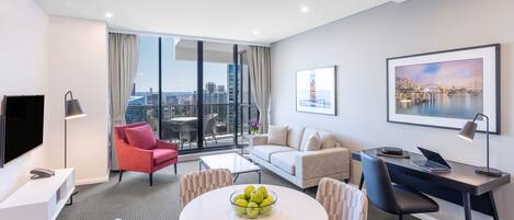 Una habitación de hotel moderna con un ventanal grande, una mesa redonda de comedor con frutero, un sofá, un escritorio con una computadora portátil y un televisor.