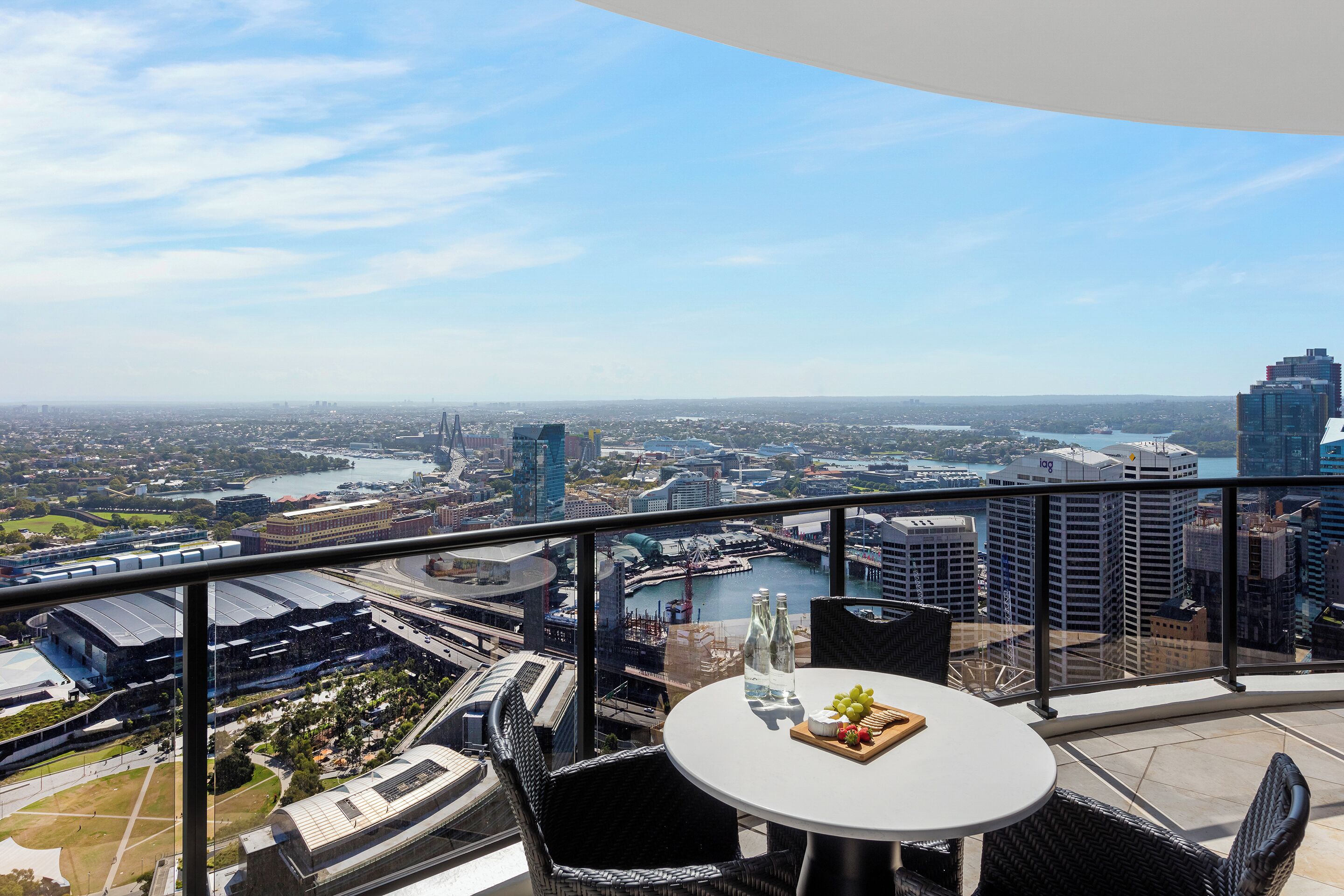 A balcony with a table, chairs, and bottles, overlooking a cityscape with buildings and a body of water.