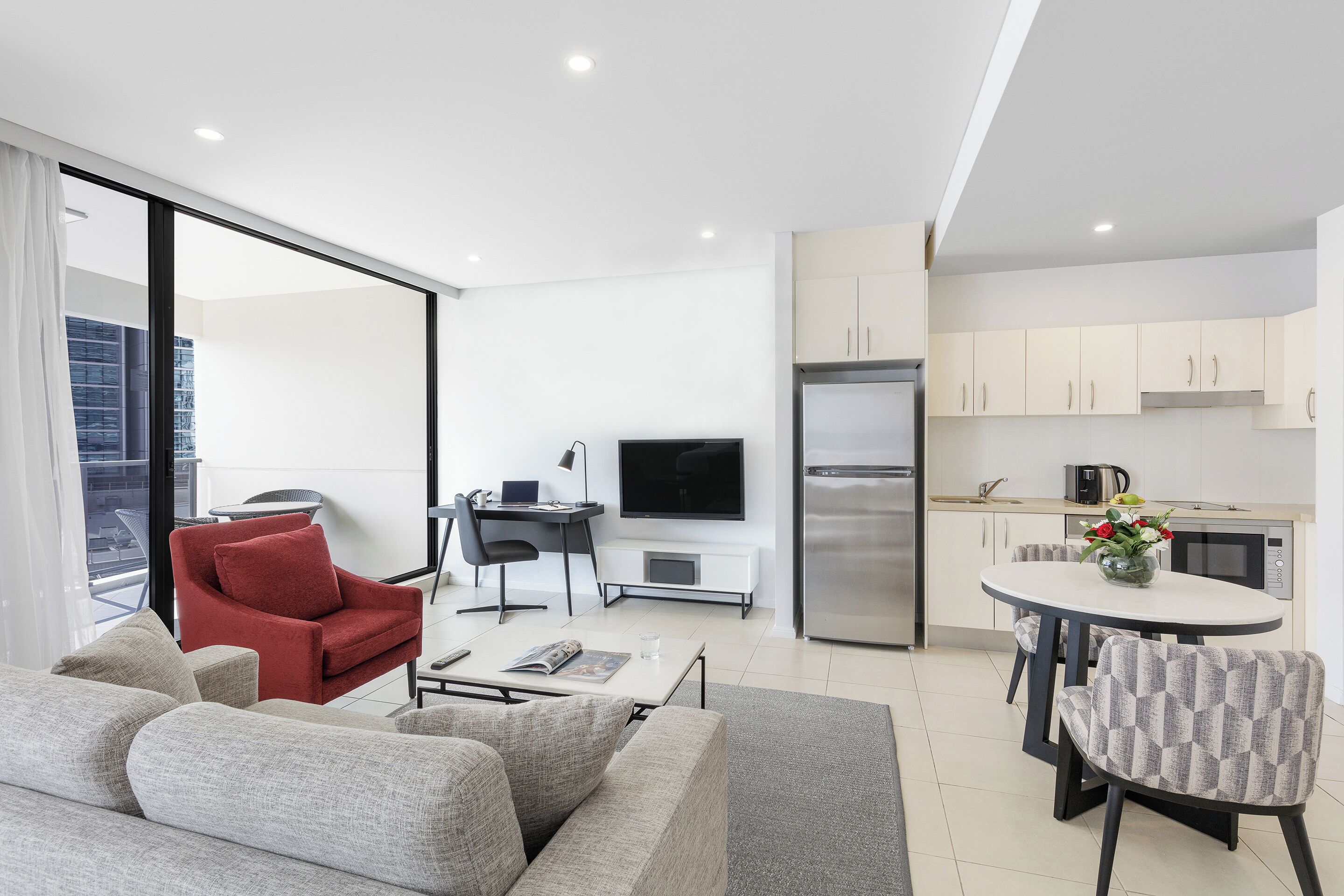 A modern living room with a sofa, a red chair, a dining table with chairs, a kitchen area with cabinets and appliances, and a large window.