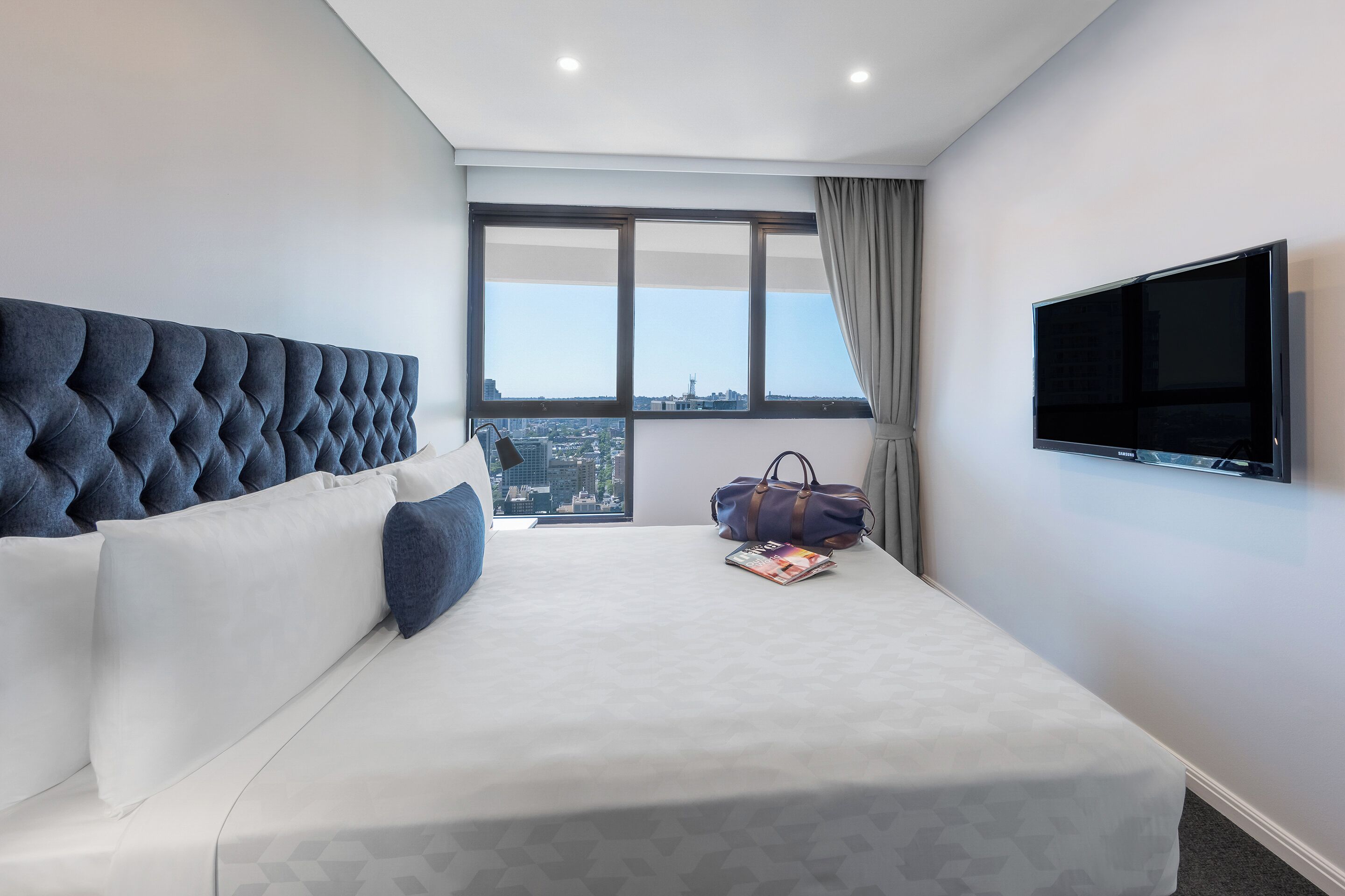 A modern hotel room with a large bed, a television, and a view of the city through a large window.