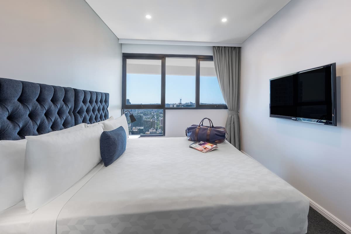 A modern hotel room with a large bed, a television, and a view of the city through a large window.