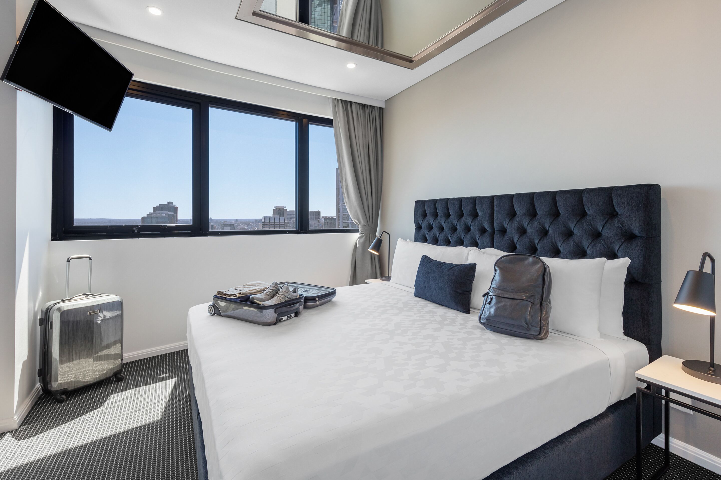 Una habitación de hotel con una cama grande, una maleta, una mochila y un televisor montado en la pared.