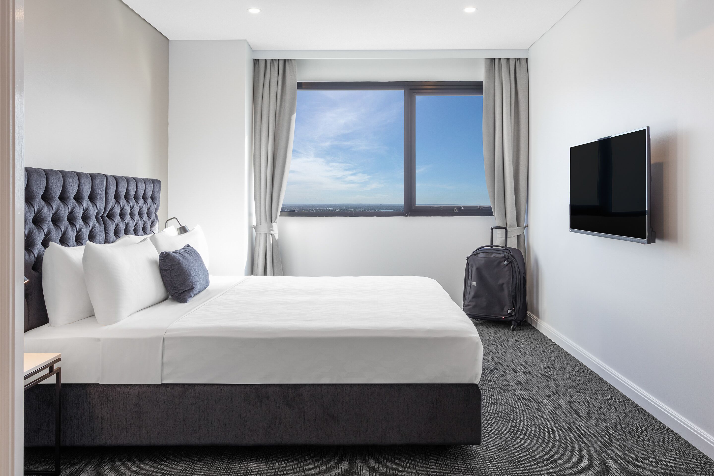 A hotel room with a large bed, a flat-screen TV, a suitcase, and a view of the city through the window.