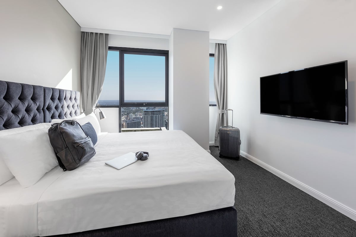 A hotel room with a large bed, a flat-screen TV, a suitcase, and a view of the city.
