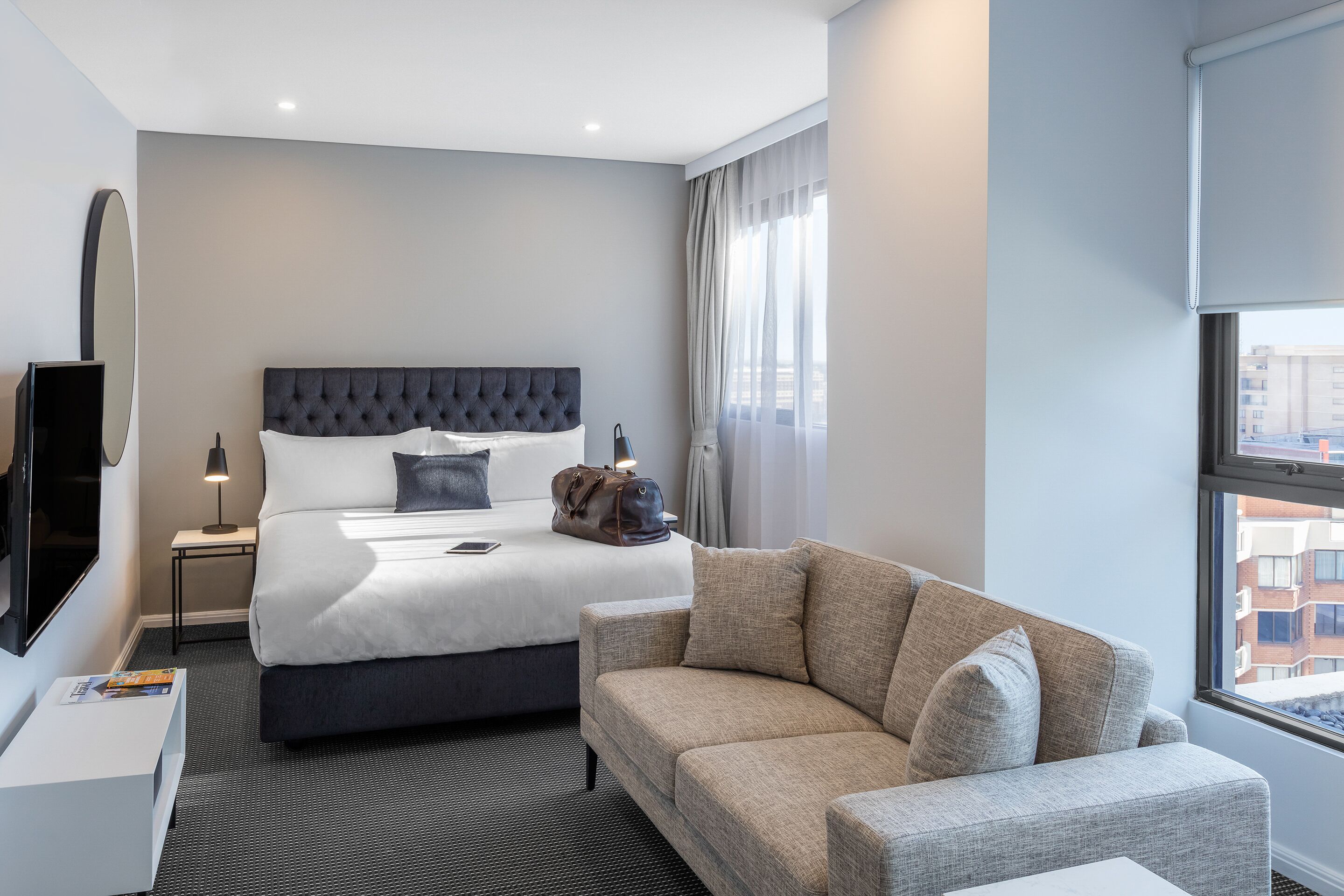Une chambre d’hôtel avec un lit, un canapé, une télévision, une lampe et une fenêtre donnant sur des immeubles.