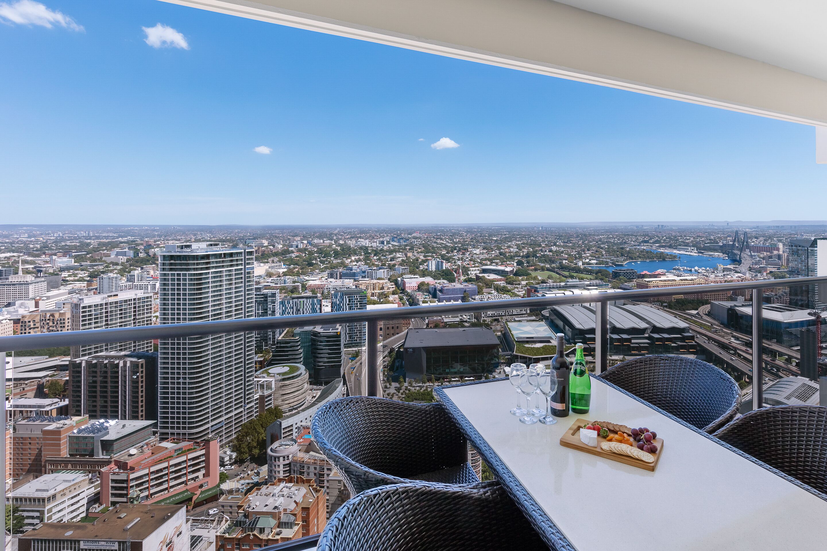 A balcony with wicker chairs and a table, overlooking a cityscape with buildings and a bridge.