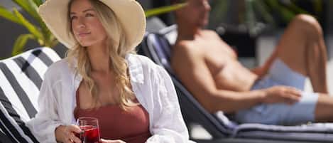 A woman in a white shirt and straw hat holding a glass of red drink, sitting on a striped lounge chair. A man is in the background on another lounge chair.