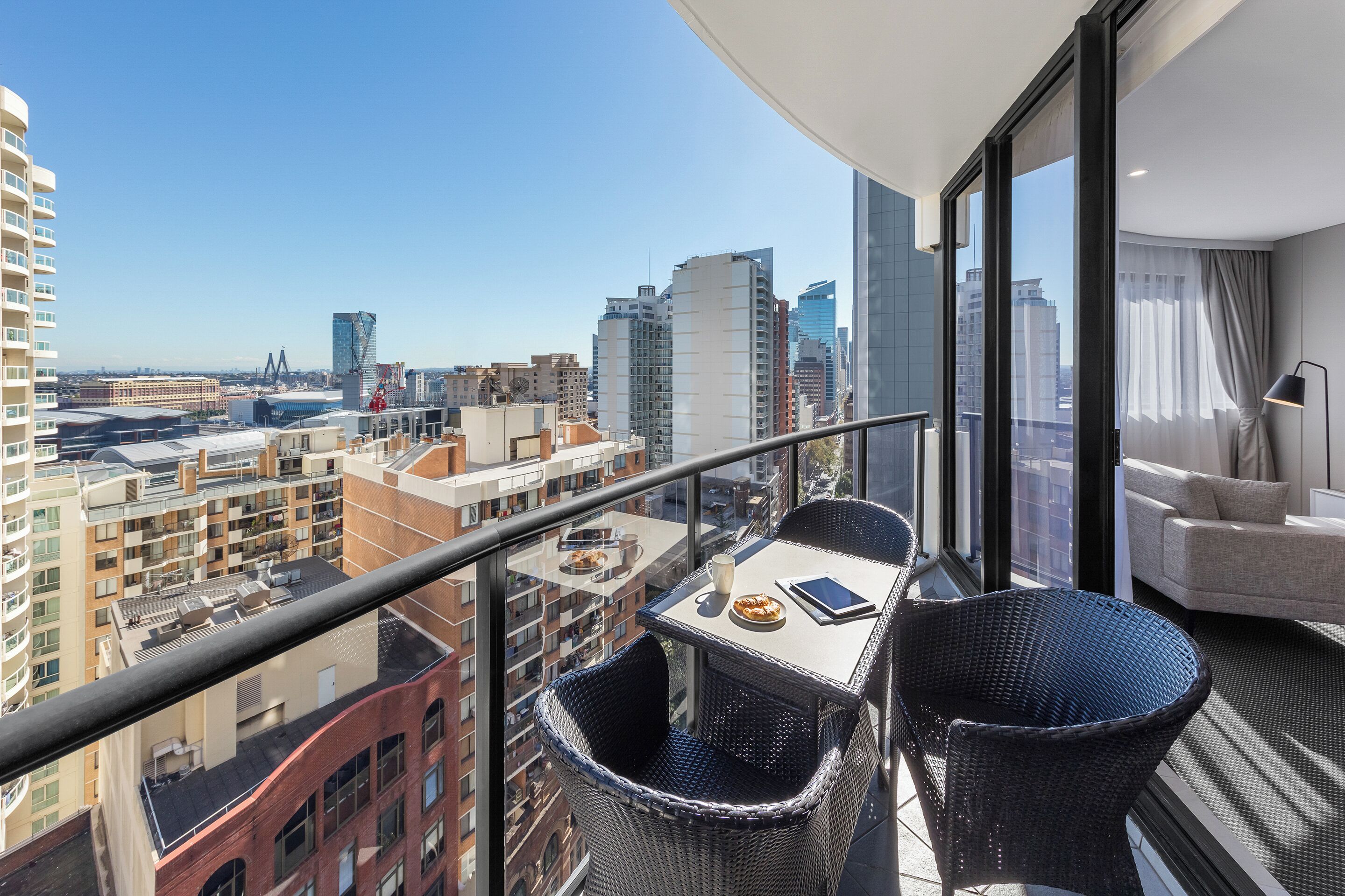 A balcony with a table, chairs, and a sofa, overlooking a cityscape with buildings and a bridge.