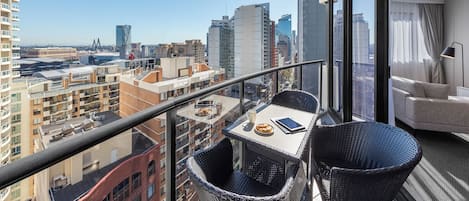 A balcony with a table, chairs, and a sofa, overlooking a cityscape with buildings and a bridge.