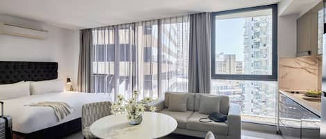 Une chambre d’hôtel moderne dotée d’un grand lit, d’un canapé, d’une table à manger ronde et offrant une vue sur le paysage urbain grâce à de grandes fenêtres.
