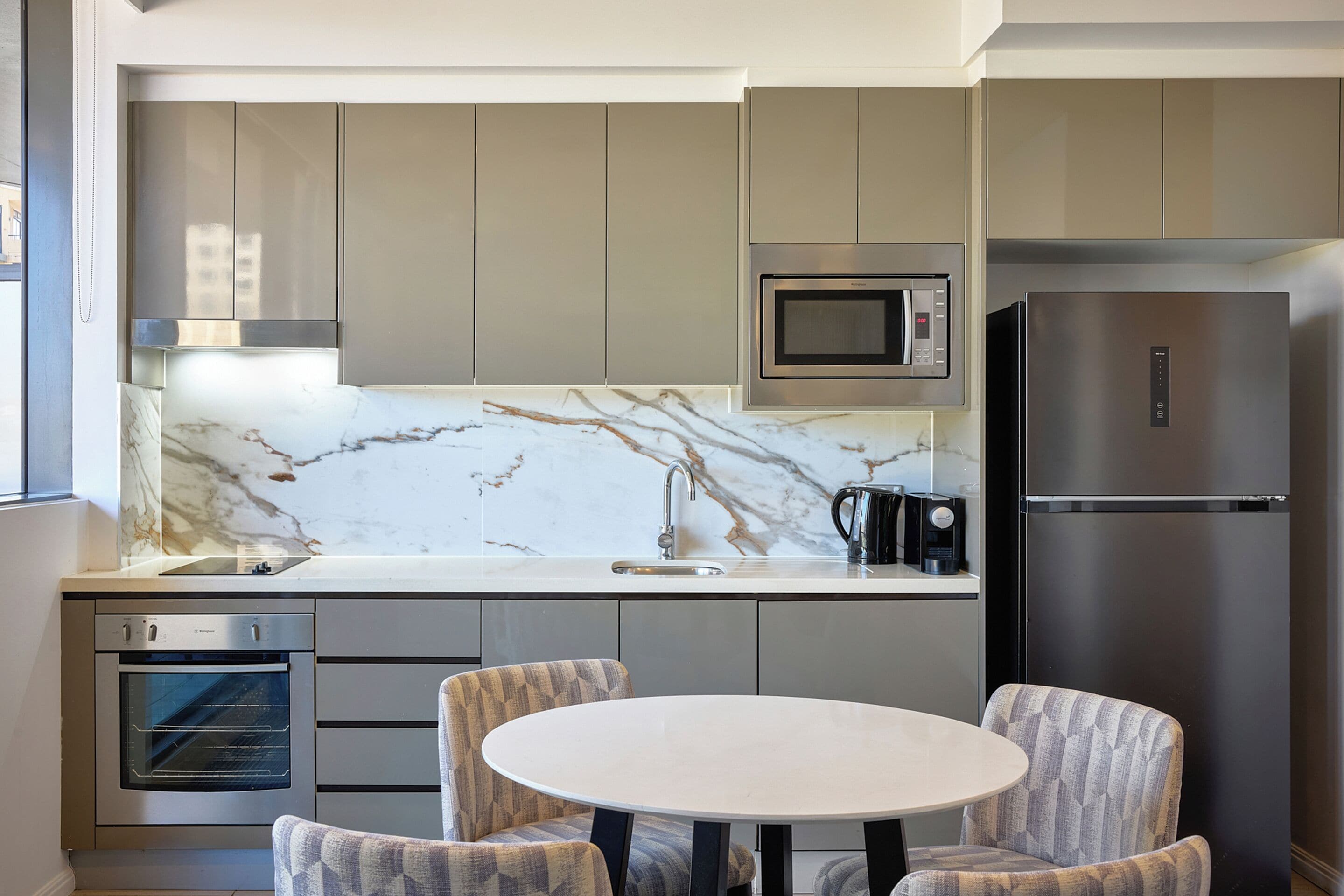 A modern kitchen with a built-in oven, microwave, and refrigerator, a marble backsplash, and a round table with two chairs.