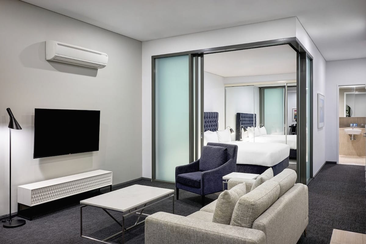 Ein modernes Hotelzimmer mit Flachbildfernseher, einer Sitzecke, einem Sessel, einem kleinen Tisch und einem Bett mit Kopfteil.