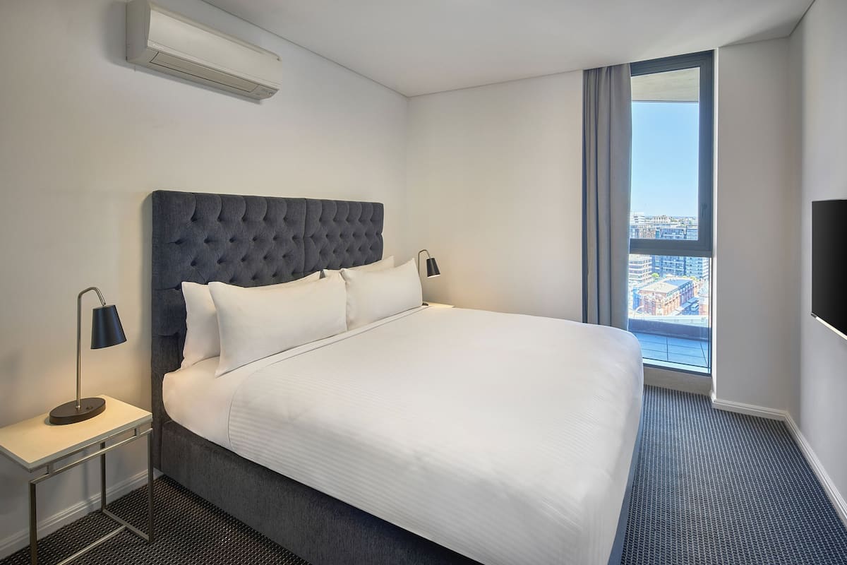 Ein Hotelzimmer mit einem großen Bett, einem Nachttisch mit Lampe, Blick auf die Stadt durch ein Fenster und einer Klimaanlage an der Wand.