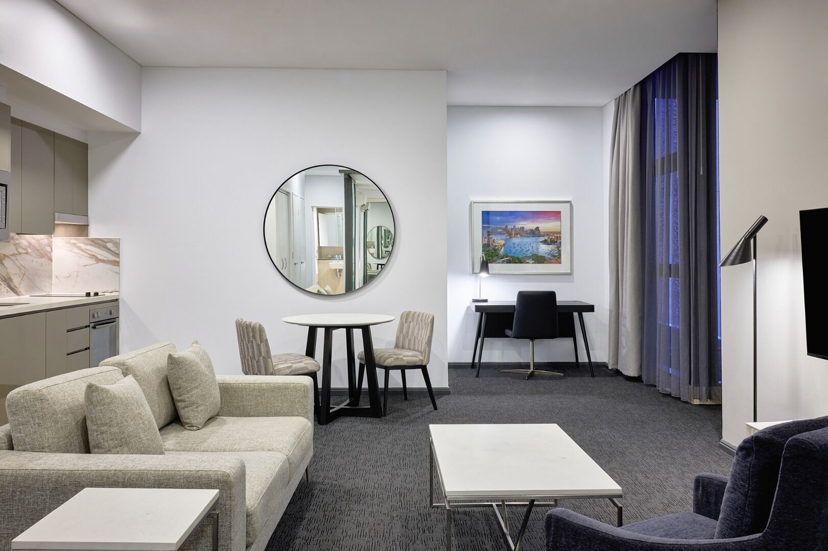 A modern hotel room with a sofa, a round mirror, a dining table with chairs, a desk, a flat-screen TV, and a painting on the wall.