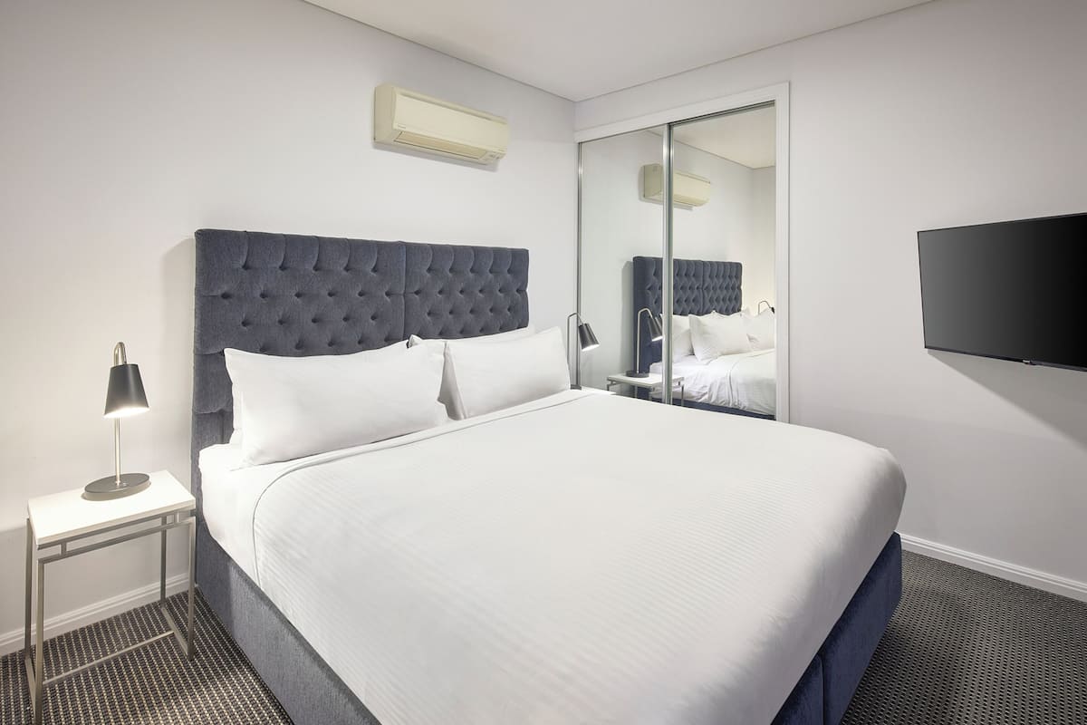 Ein Hotelzimmer mit einem großen Bett, einem Nachttisch mit Lampe, einem Flachbildfernseher und einer Schiebetür zum Kleiderschrank.