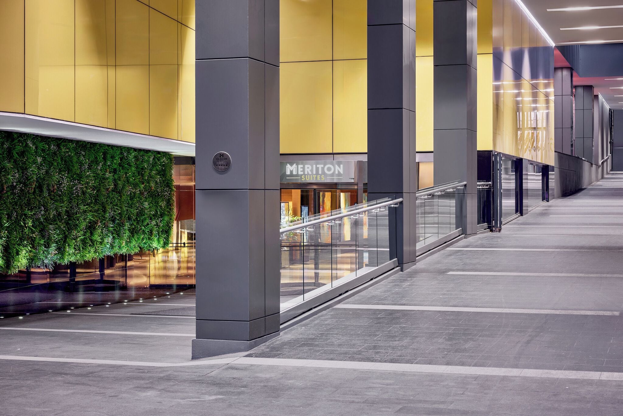 A modern hotel corridor with a glass railing, a green wall, and a sign for Meriton Suites.
