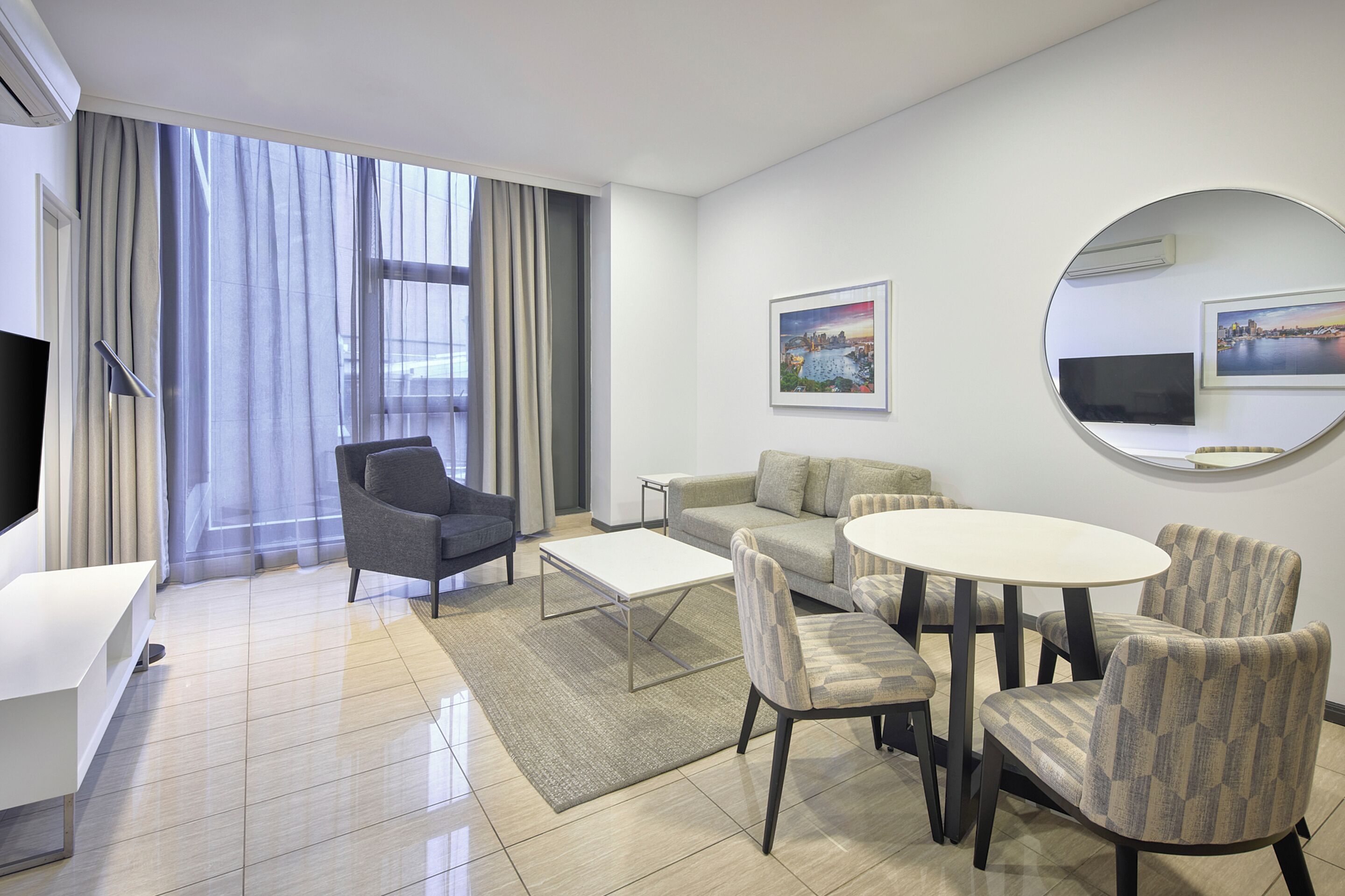 A modern hotel room with a sofa, armchairs, a small table, a TV, and a large window with curtains.