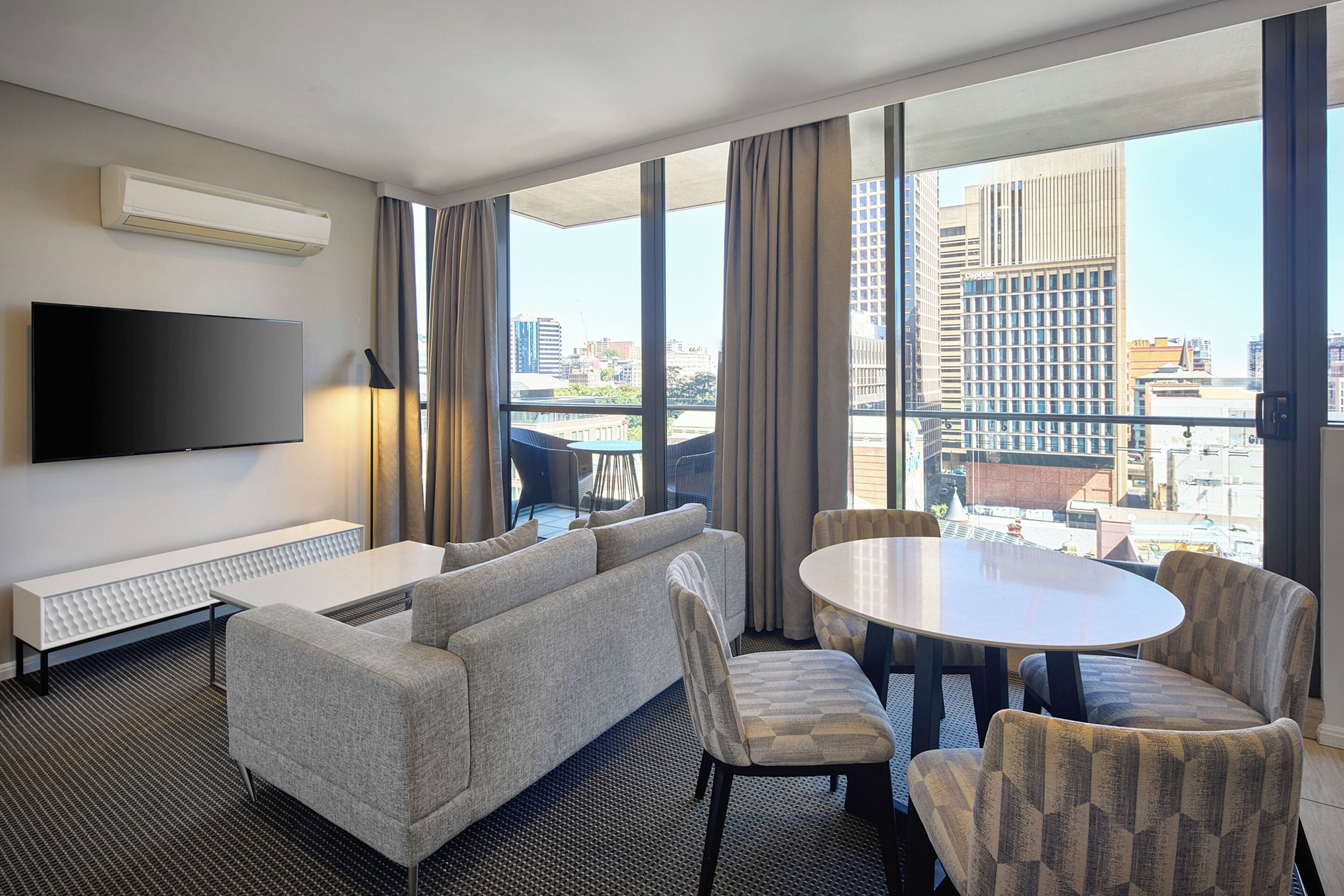 A hotel room with a large window, a flat-screen TV, a sofa, a dining table, and chairs.