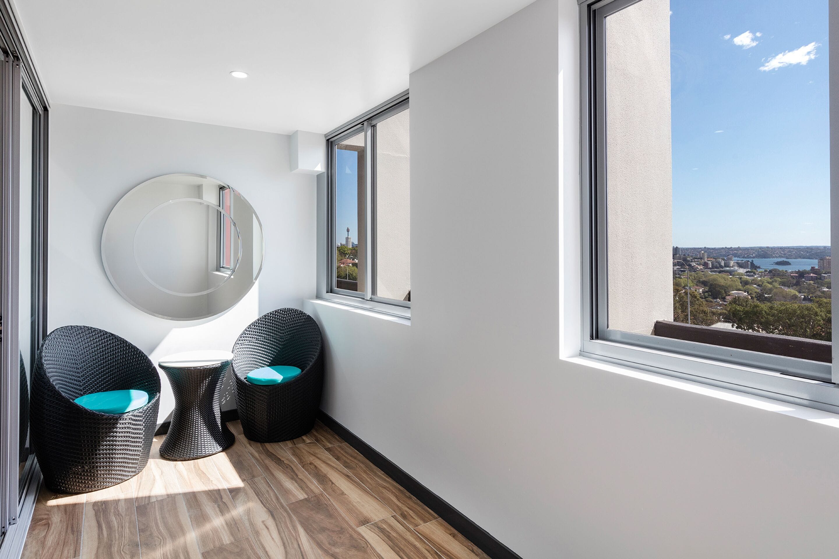 Ein modernes Zimmer mit einem großen Fenster, einem runden Spiegel und zwei Korbstühlen mit türkisfarbenen Kissen.