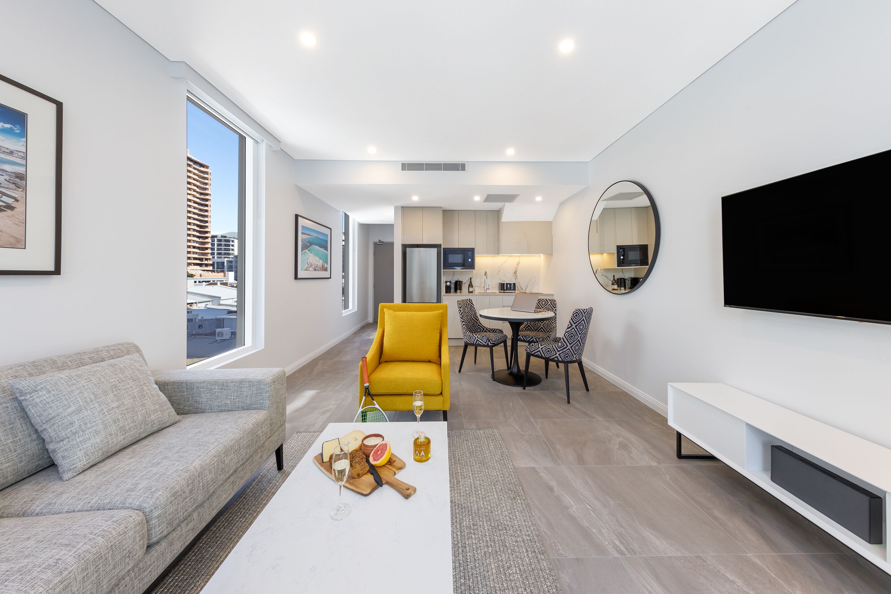 A modern living room with a sofa, a coffee table with food, a yellow armchair, a dining area with chairs, a kitchen with appliances, and a large TV.