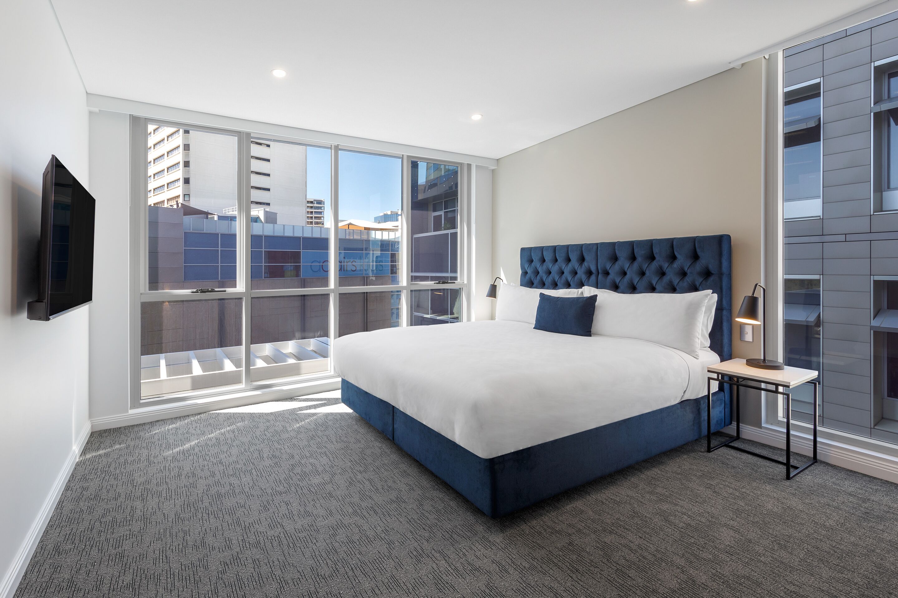 A modern hotel bedroom with a large bed, a bedside table with a lamp, and a view of the city through floor-to-ceiling windows.