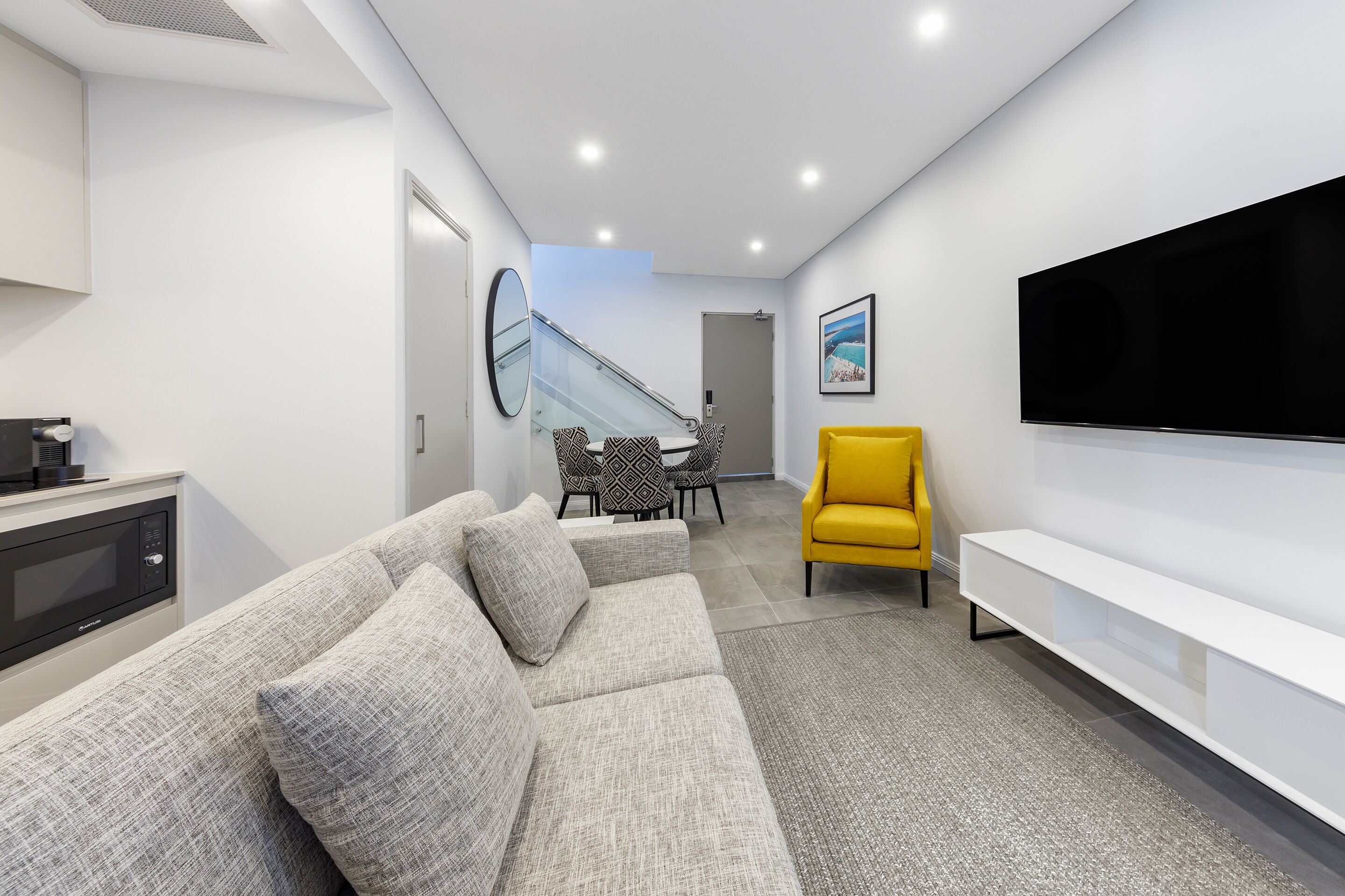 Una sala de estar moderna con un sofá gris, un sillón amarillo, un televisor de pantalla plana y un comedor con sillas estampadas.