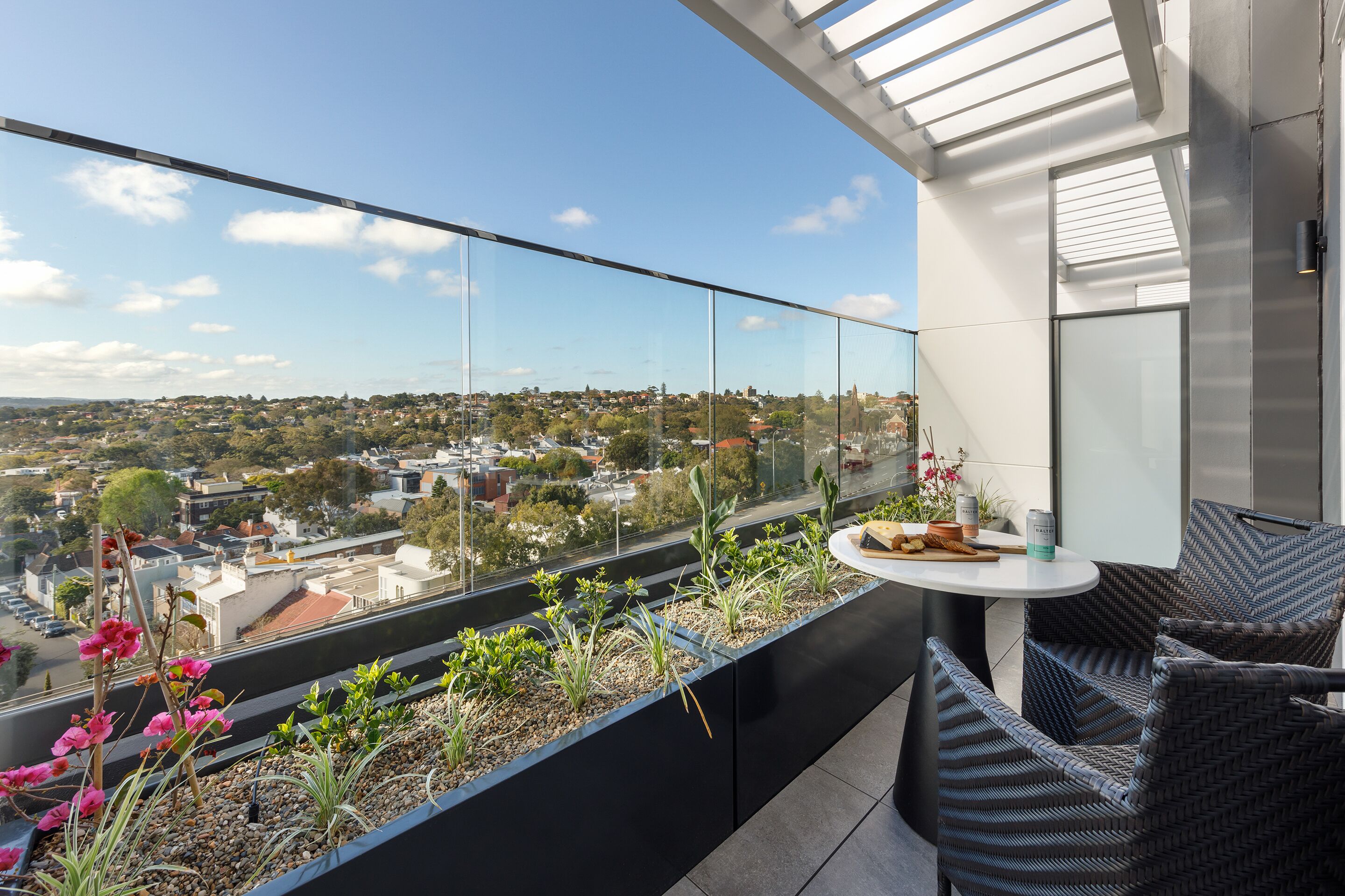 Een balkon met glazen hekwerk, een tafel met stoelen en uitzicht op een stadsbeeld.