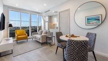 A modern living room with a sofa, yellow armchair, patterned chairs, a round table, and a large window offering a city view.