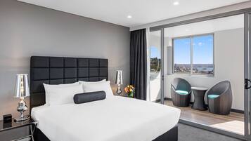 A modern hotel room with a large bed, two bedside lamps, a view of the city, and a balcony with wicker chairs.