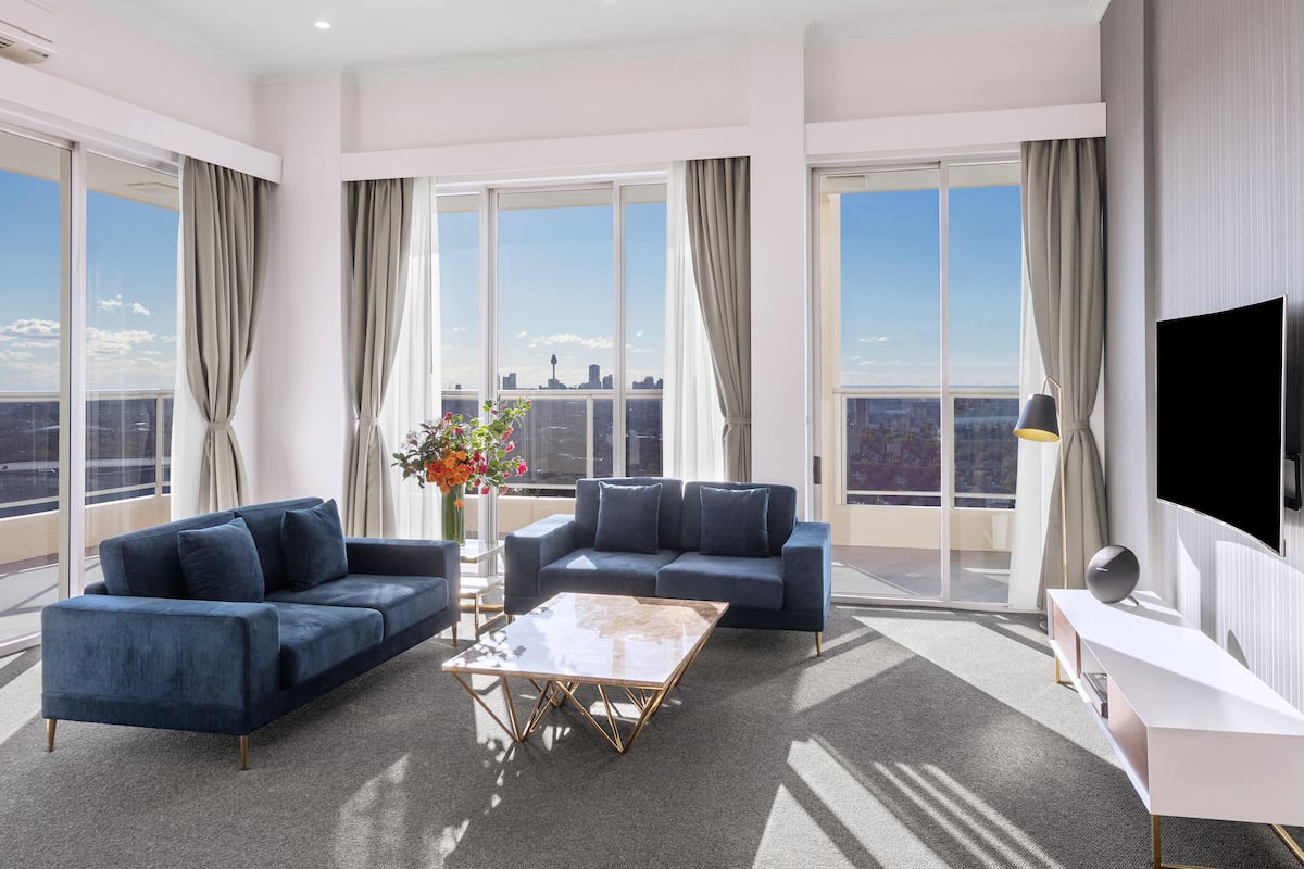 Un salon moderne avec deux canapés bleus, une table basse et un téléviseur. De grandes fenêtres offrent une vue sur la ville.