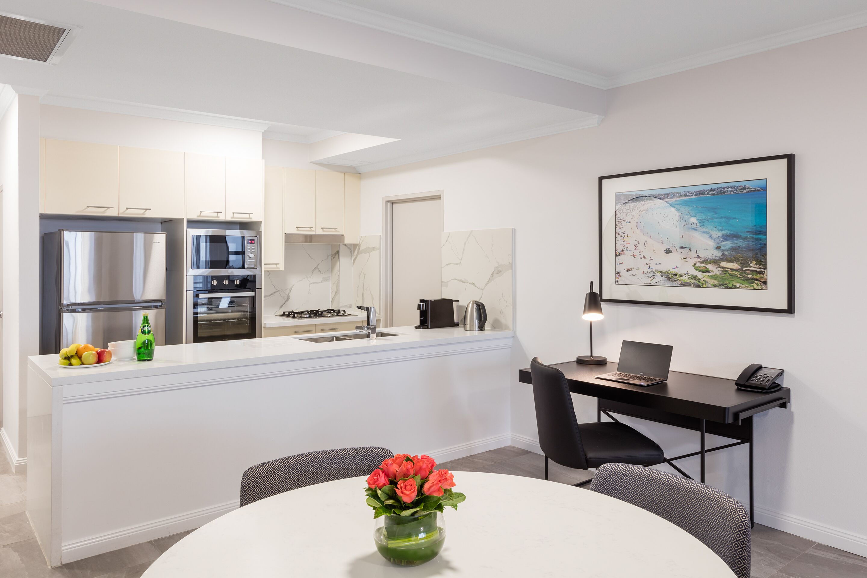 A modern kitchen with white cabinets, a stainless steel refrigerator, and a microwave. There's a dining area with a round table and a vase of flowers. A framed beach painting is on the wall.