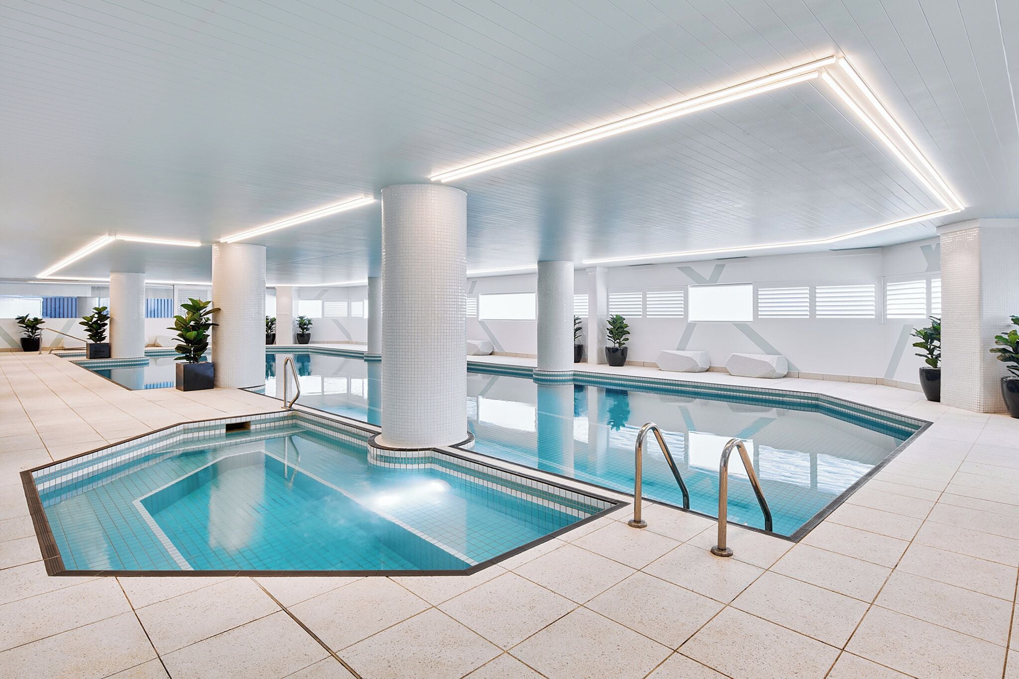 Une piscine intérieure moderne, avec une eau d’un bleu limpide, entourée de sols et de murs recouverts de carreaux blancs, et éclairée par des luminaires linéaires encastrés au plafond.