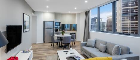 A modern living room with a grey sofa, yellow armchairs, a white coffee table, and a flat-screen TV. There is a dining area with a round table and chairs, and a kitchen in the background.