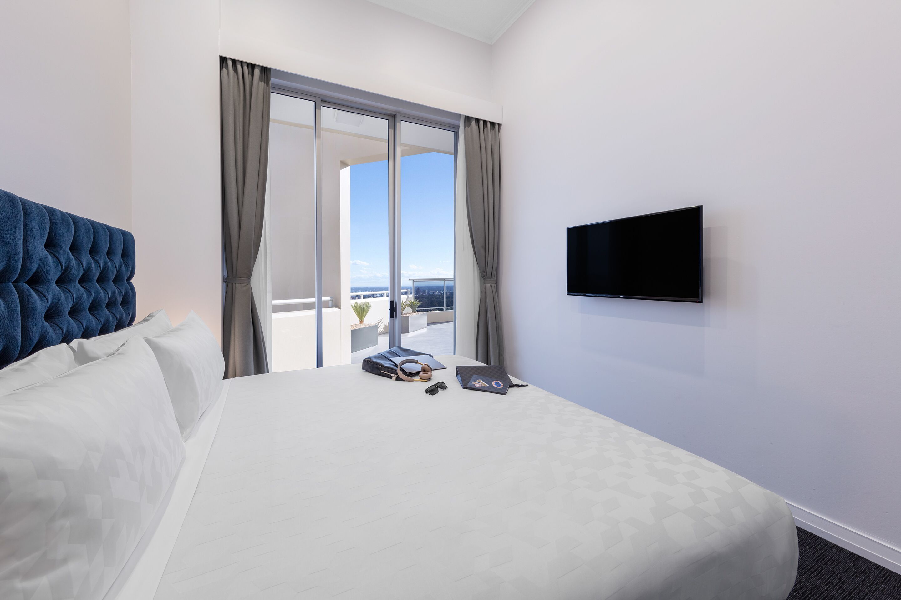 A hotel room with a large bed, a flat-screen TV mounted on the wall, and a view of the city through a sliding glass door.