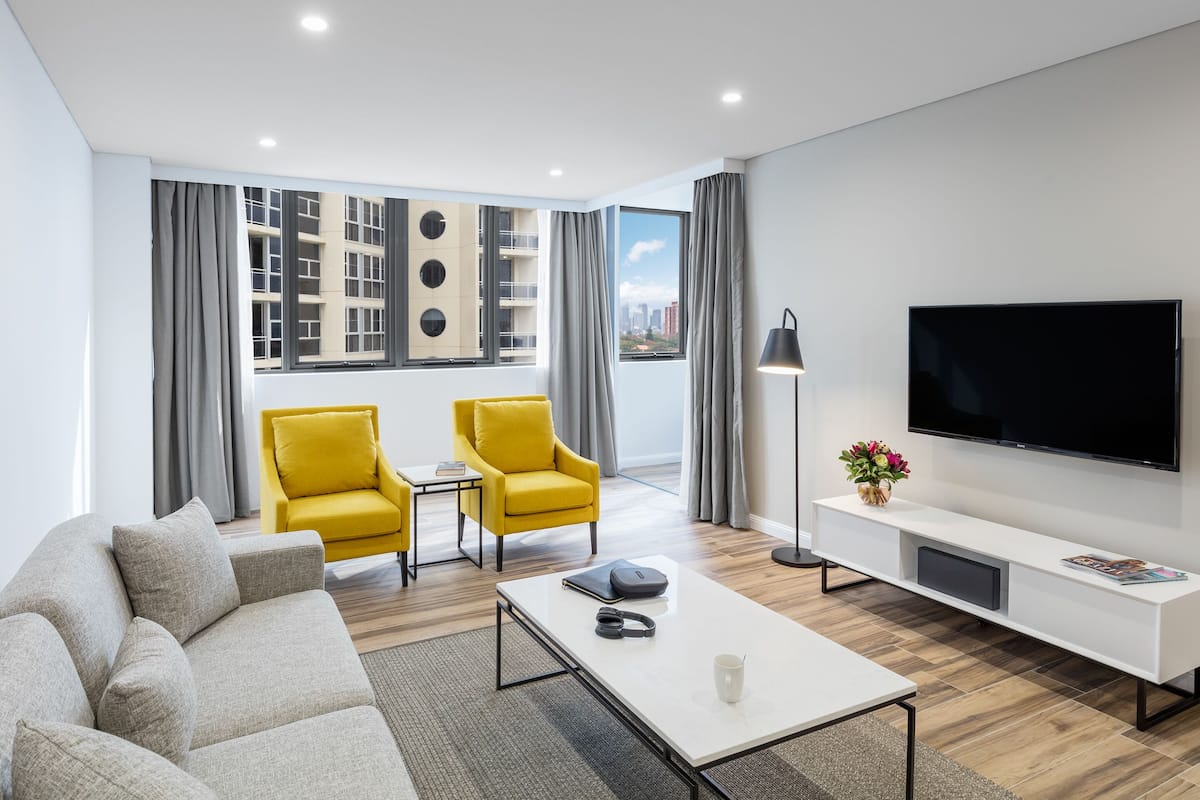 Un salon moderne avec un canapé, deux fauteuils jaunes, une table basse, un téléviseur à écran plat et une grande fenêtre avec des rideaux.
