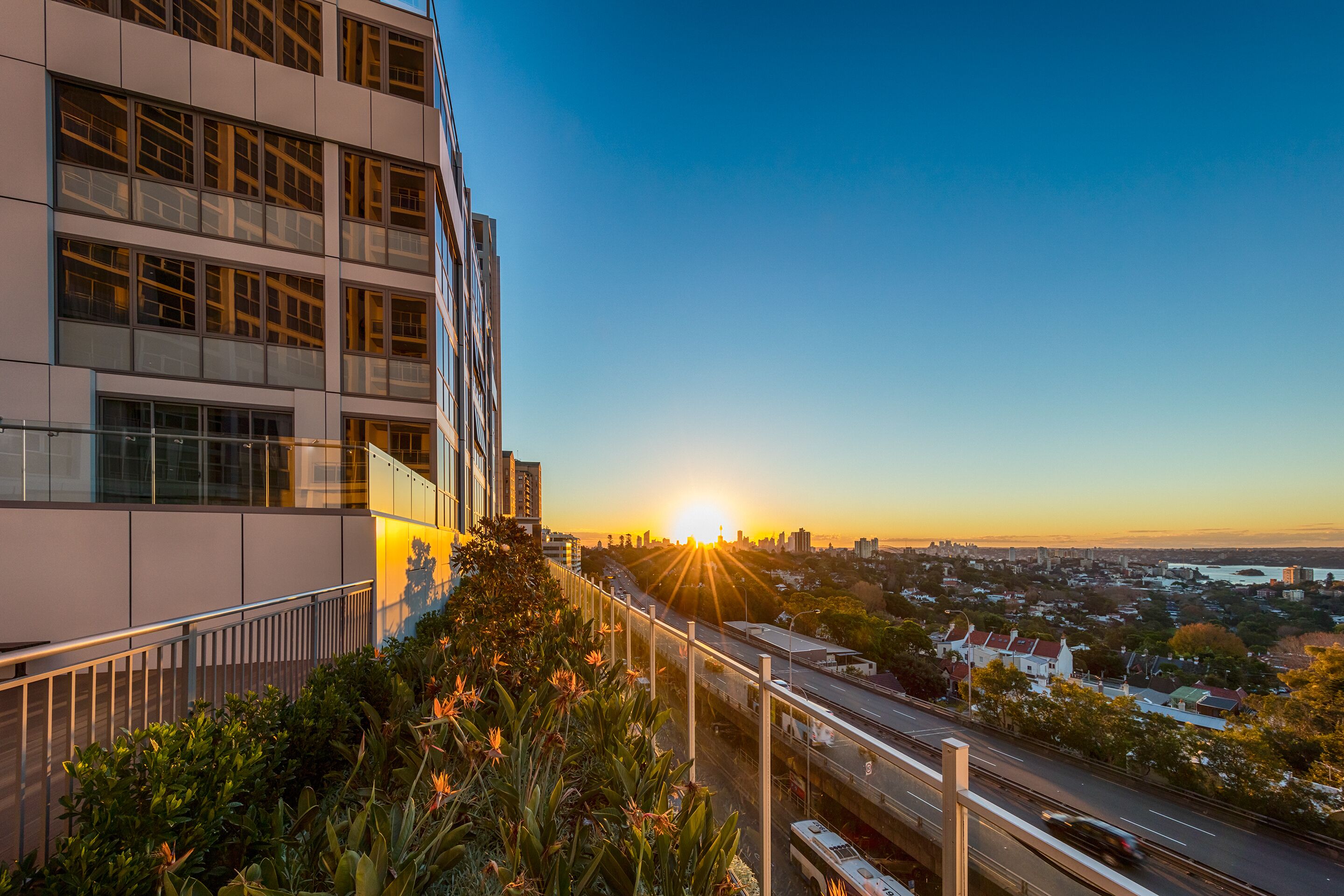 A modern building with multiple floors and large windows, overlooking a busy road and a cityscape during sunset.