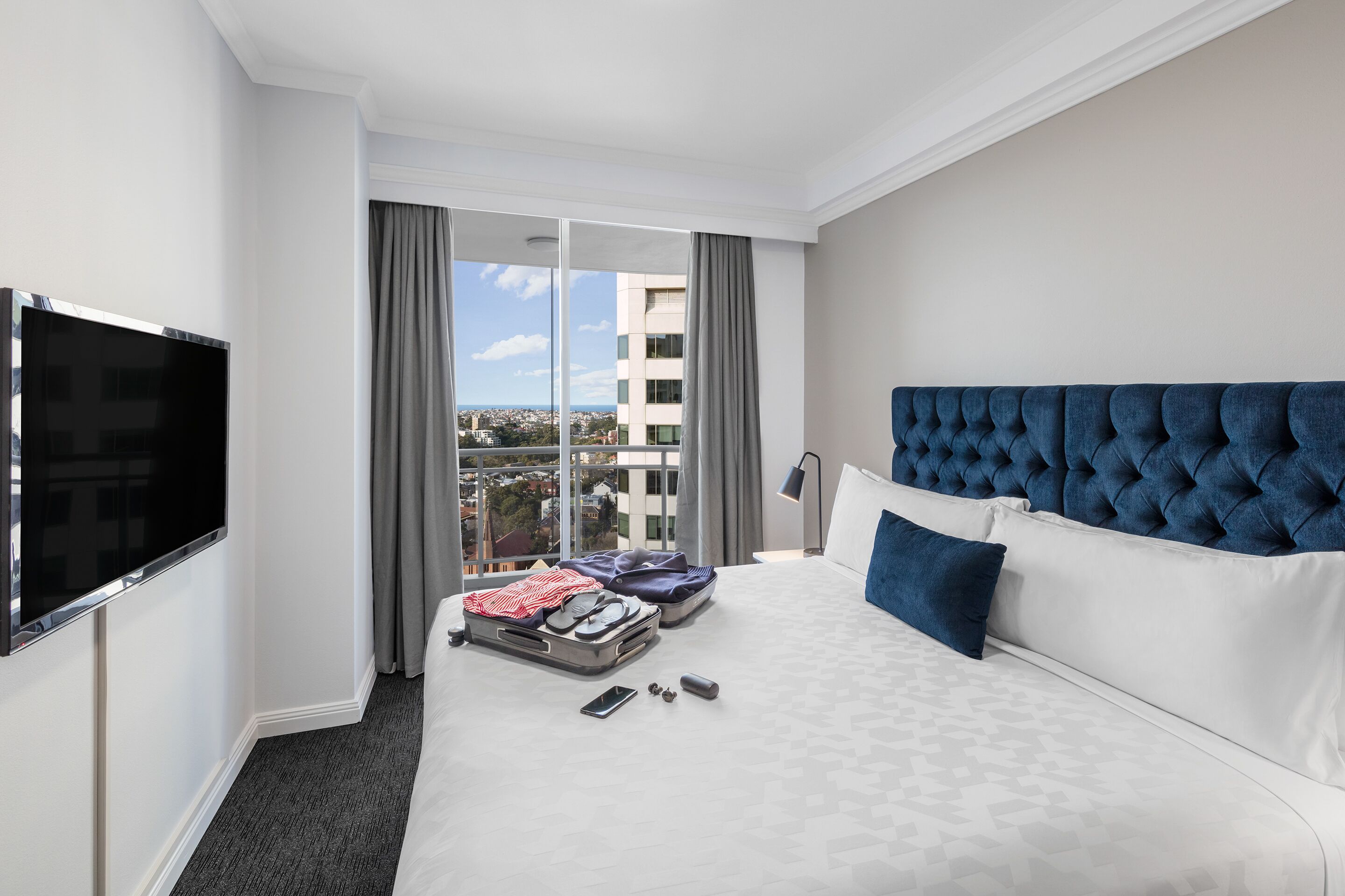 A hotel room with a large bed, a flat-screen TV, a view of the city, and a window with curtains.