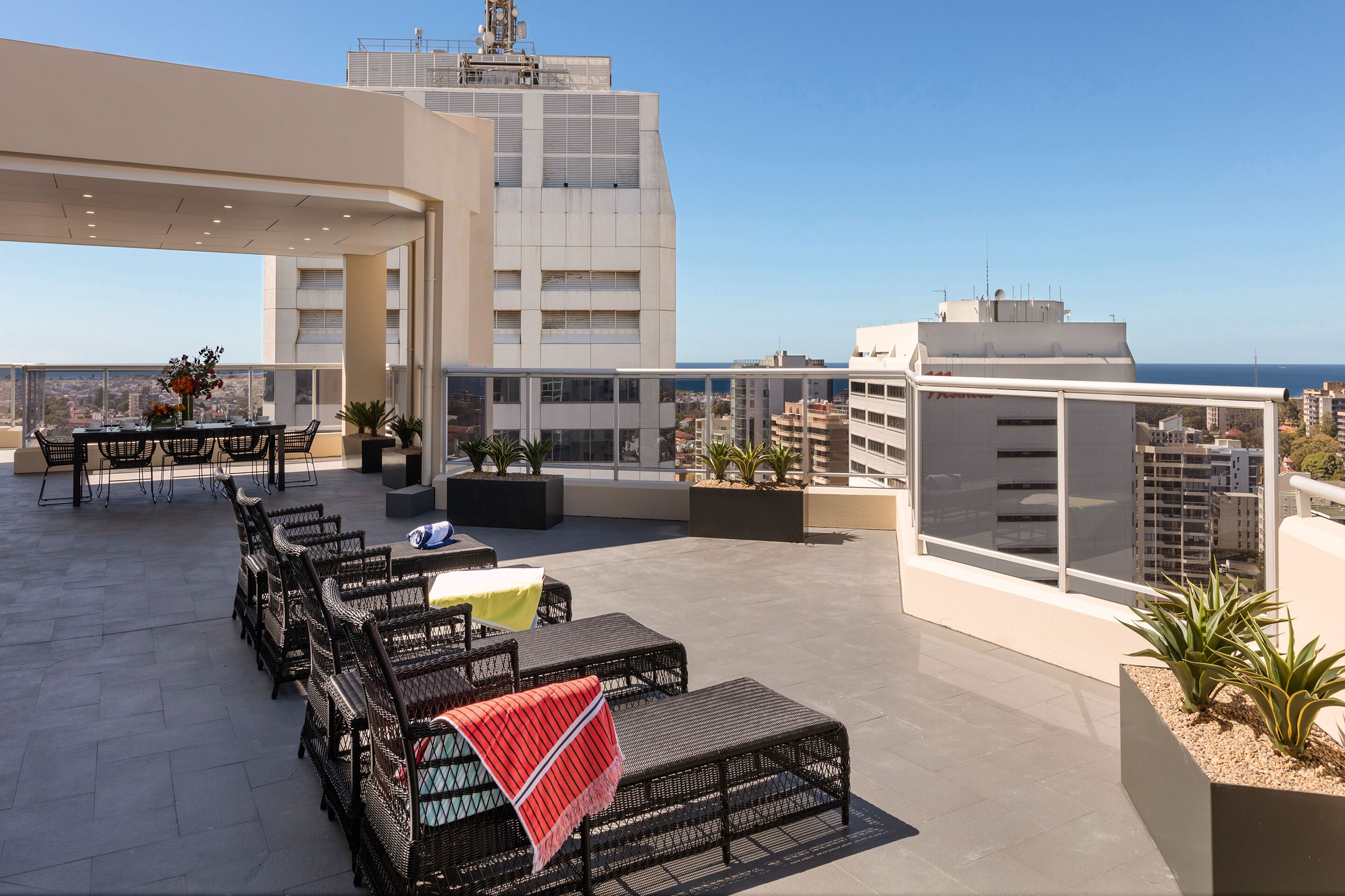 A rooftop terrace with outdoor seating, potted plants, and a view of the city and sea.